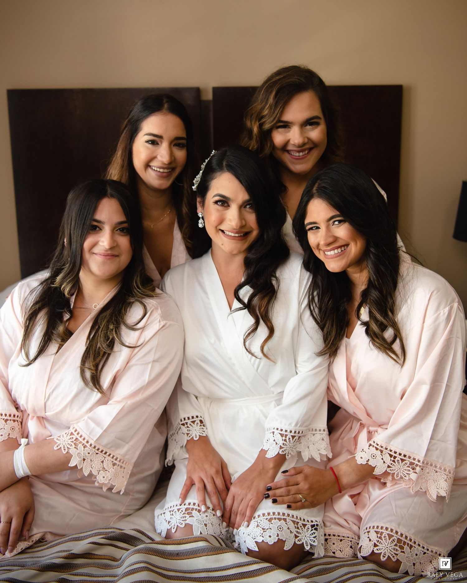 Bride in white robe poses with four bridesmaids in pink robes on bed during wedding morning preparations.