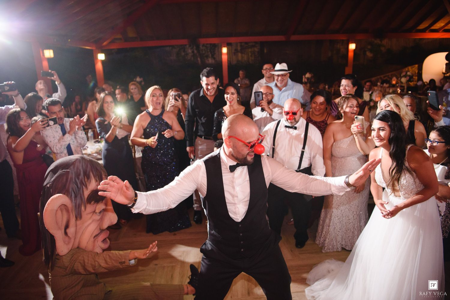 Guests dancing joyfully at an elegant wedding reception, with the bride in a white gown and a man in a tuxedo wearing red glasses.