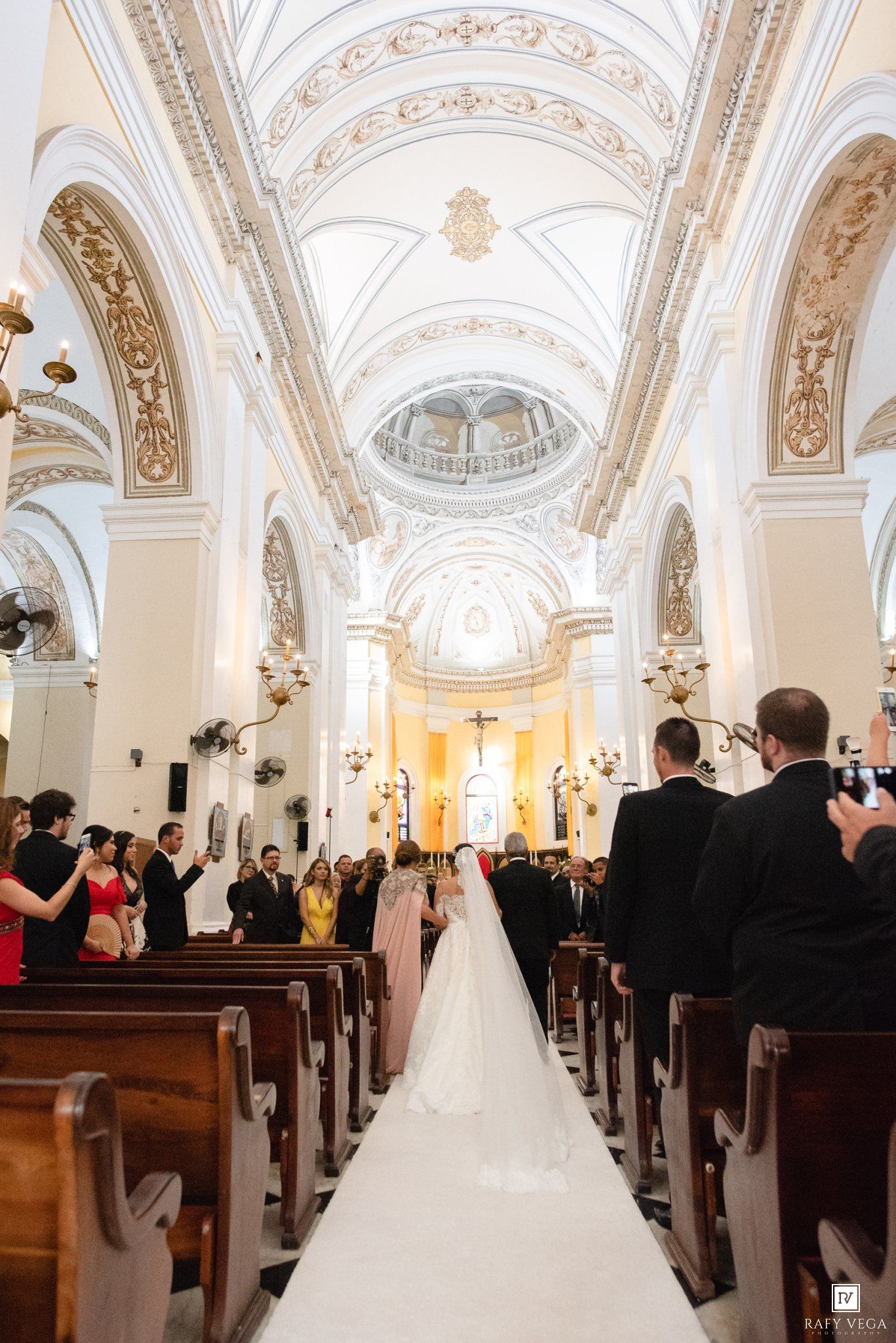 Bride walks down the aisle in a grand white church with ornate vaulted ceilings during a traditional wedding ceremony.