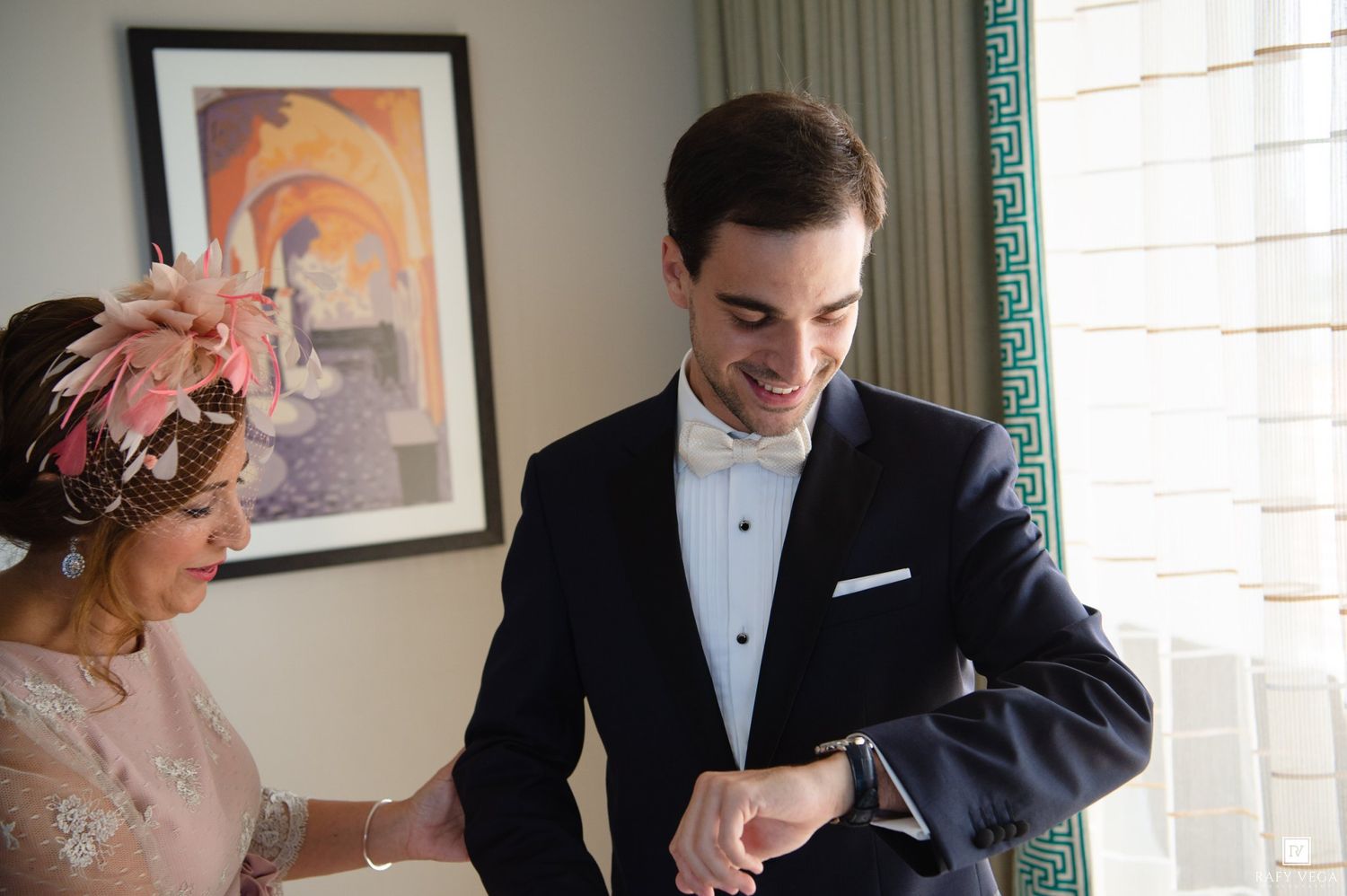 Groom in navy tuxedo checking cufflinks assisted by woman in pink lace dress at wedding preparation.