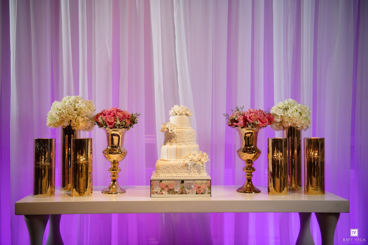Elegant wedding cake display on white table with gold vases, pink floral arrangements, and purple draped backdrop.