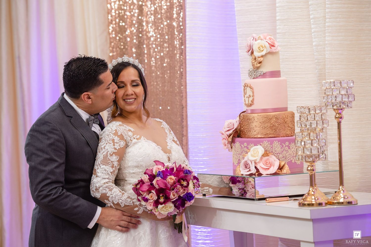 Newlywed couple sharing a tender kiss beside an elegant pink and gold tiered wedding cake at their reception.