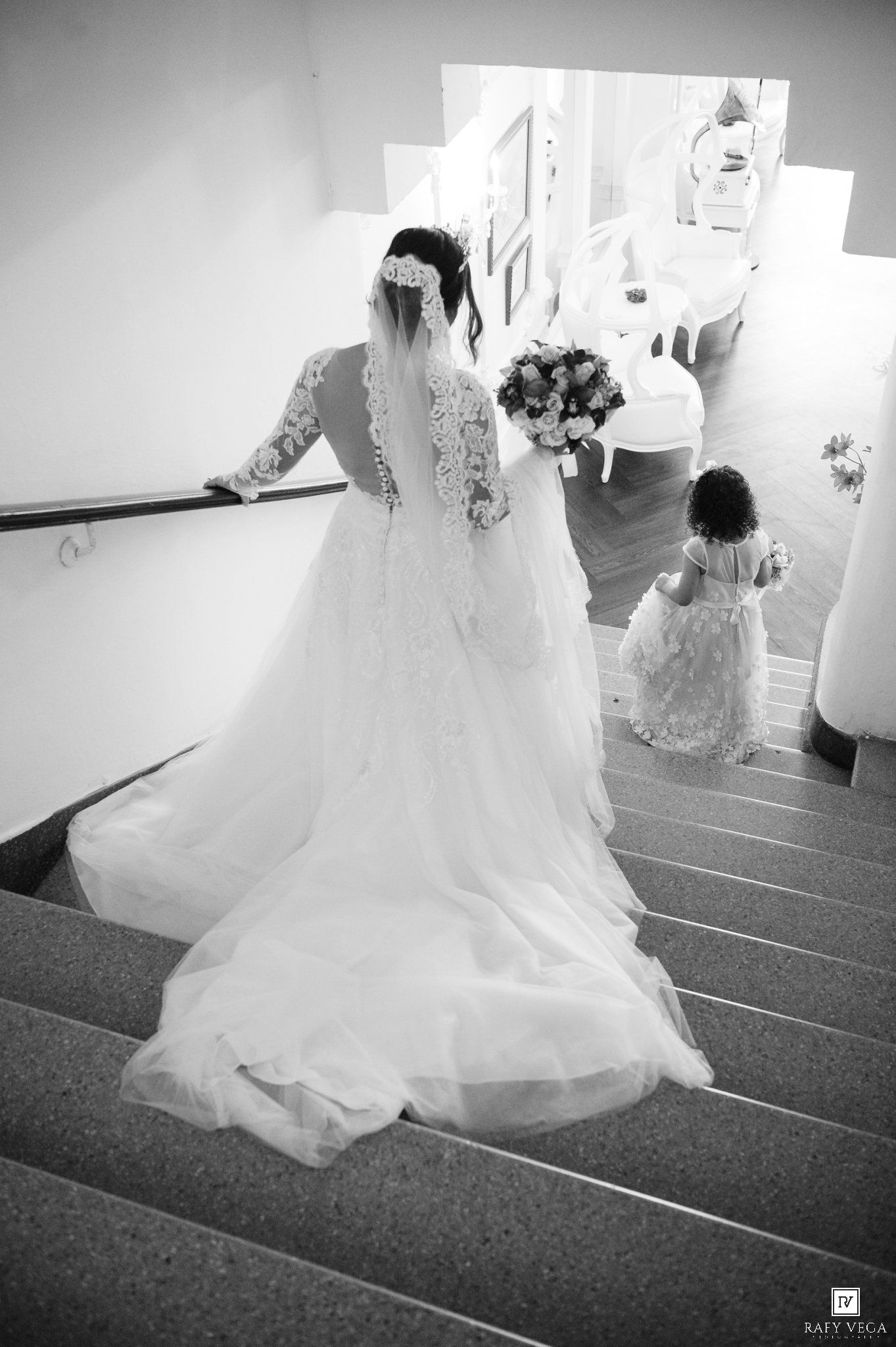 Bride in lace wedding gown with veil descends stairs with flower girl in white dress, black and white wedding photo.