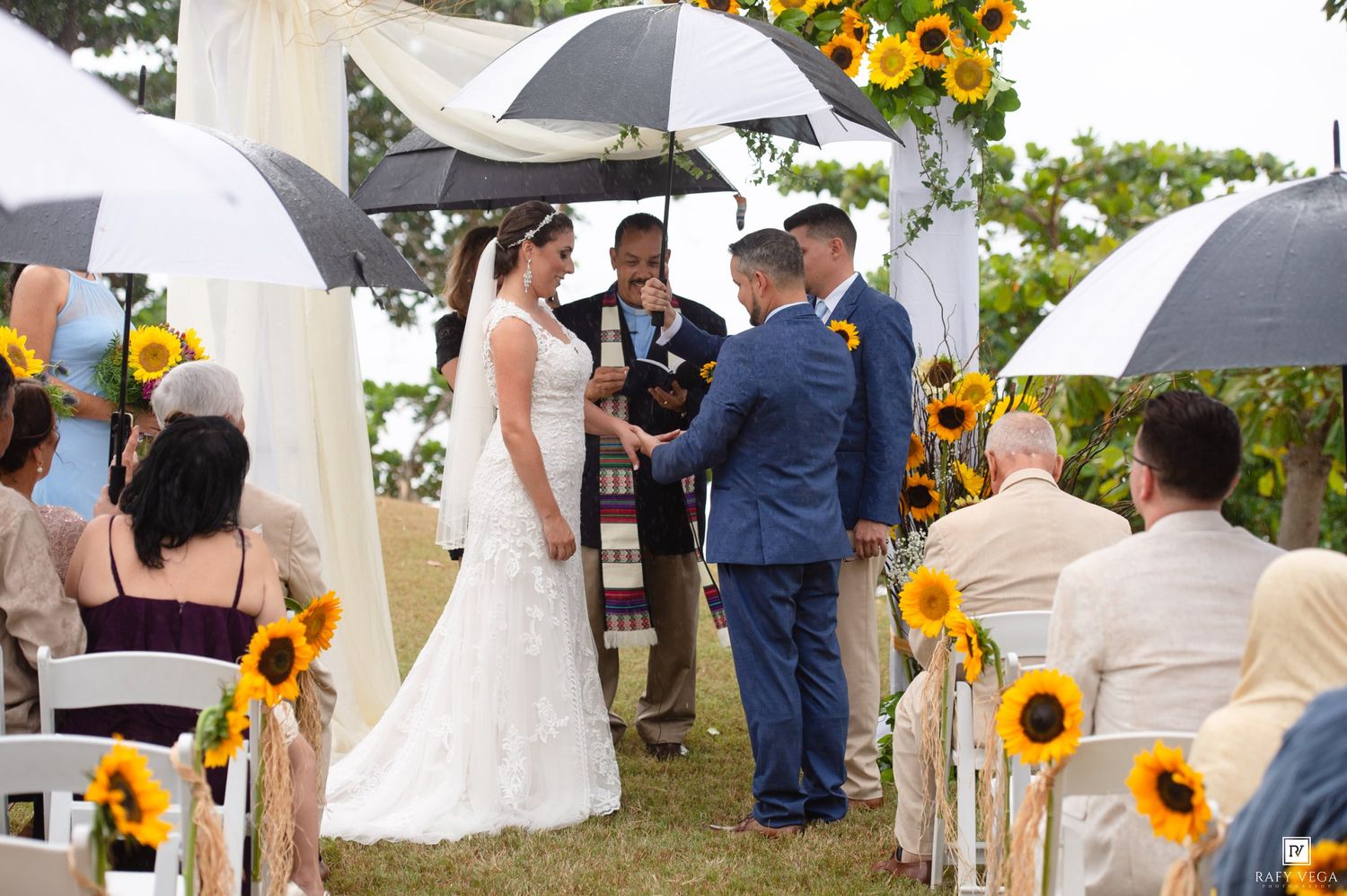 Outdoor wedding ceremony with sunflowers, couple exchanging vows under umbrellas on a cloudy day.