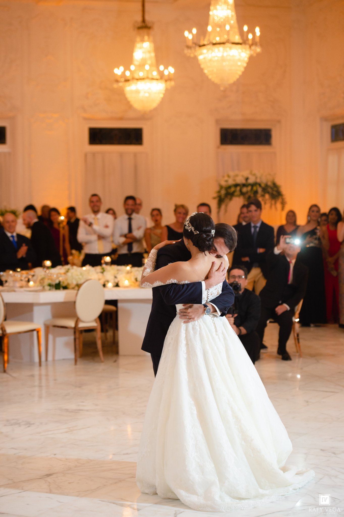 Bride and groom share romantic first dance in grand ballroom with chandeliers and guests watching.