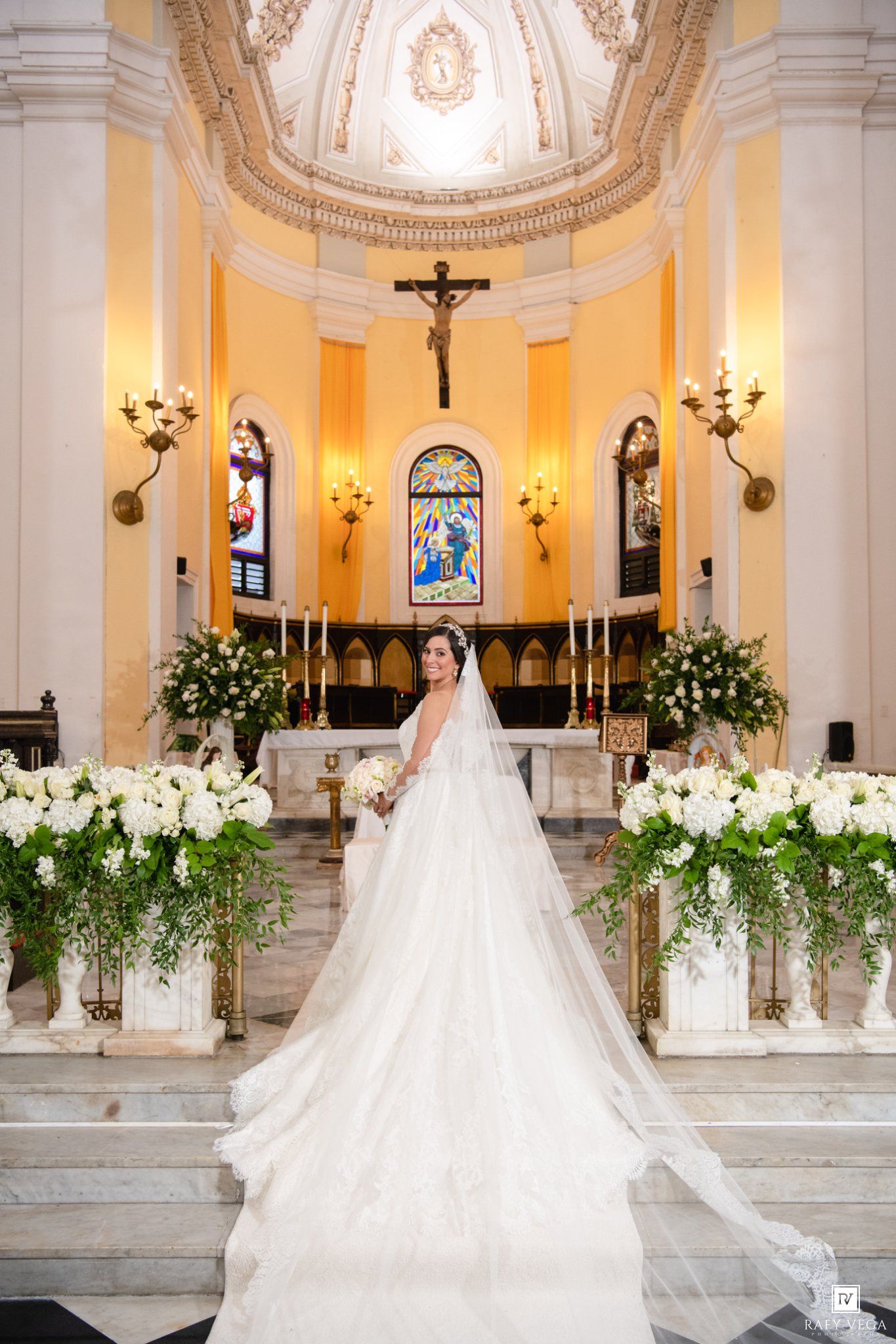 Bride in a stunning white ball gown with long veil stands at church altar adorned with white floral arrangements.