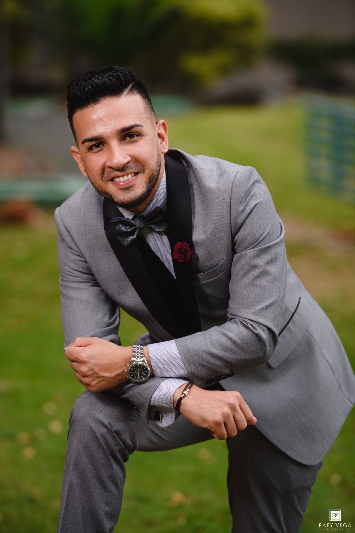 Smiling groom in gray tuxedo with black lapels and bow tie posing outdoors in a garden setting.