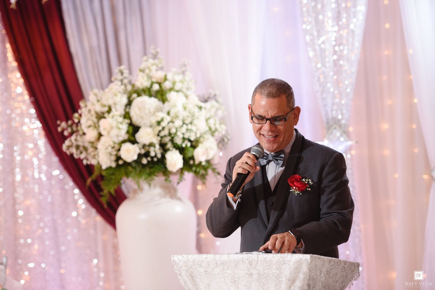 Man in suit speaking at wedding reception podium with white floral arrangement and elegant draped backdrop.