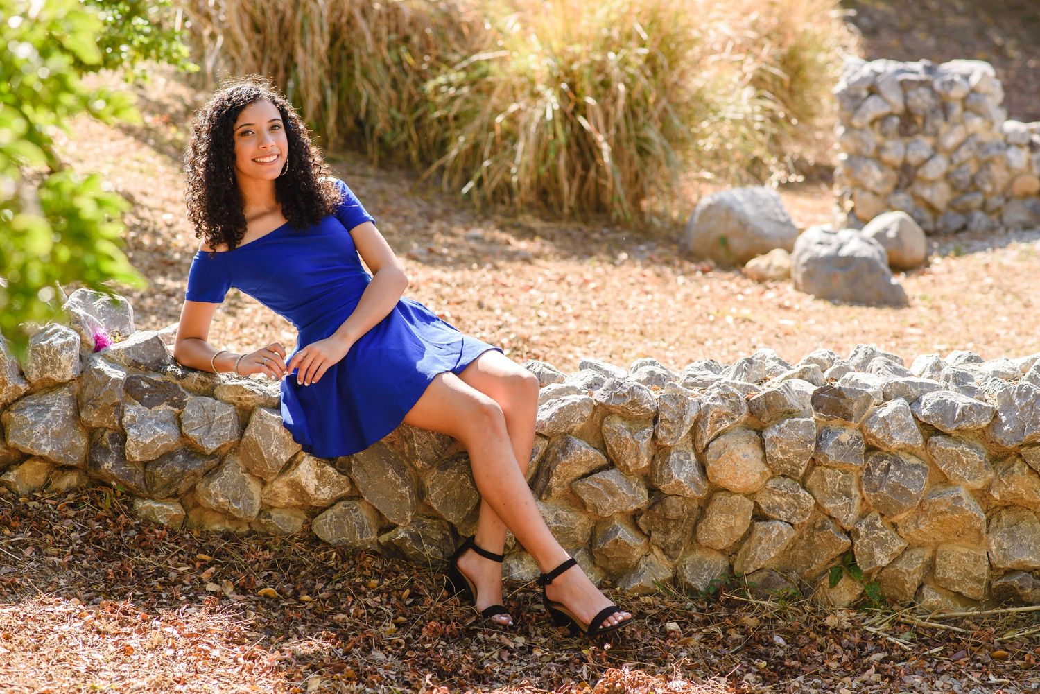 Young woman in blue dress sitting on stone wall in sunny outdoor garden setting.