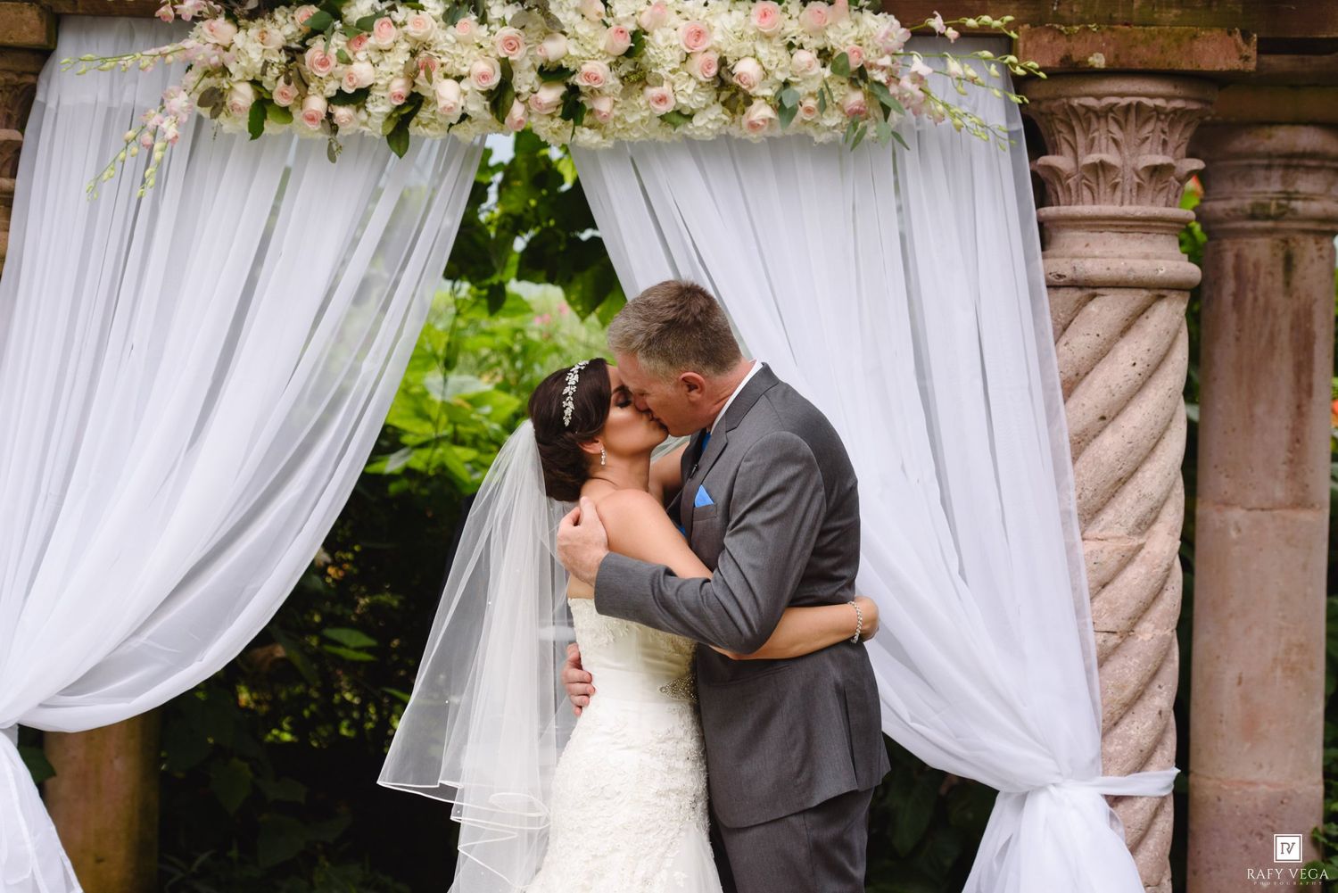 Bride and groom share first kiss under floral arch with white draping at outdoor wedding ceremony.