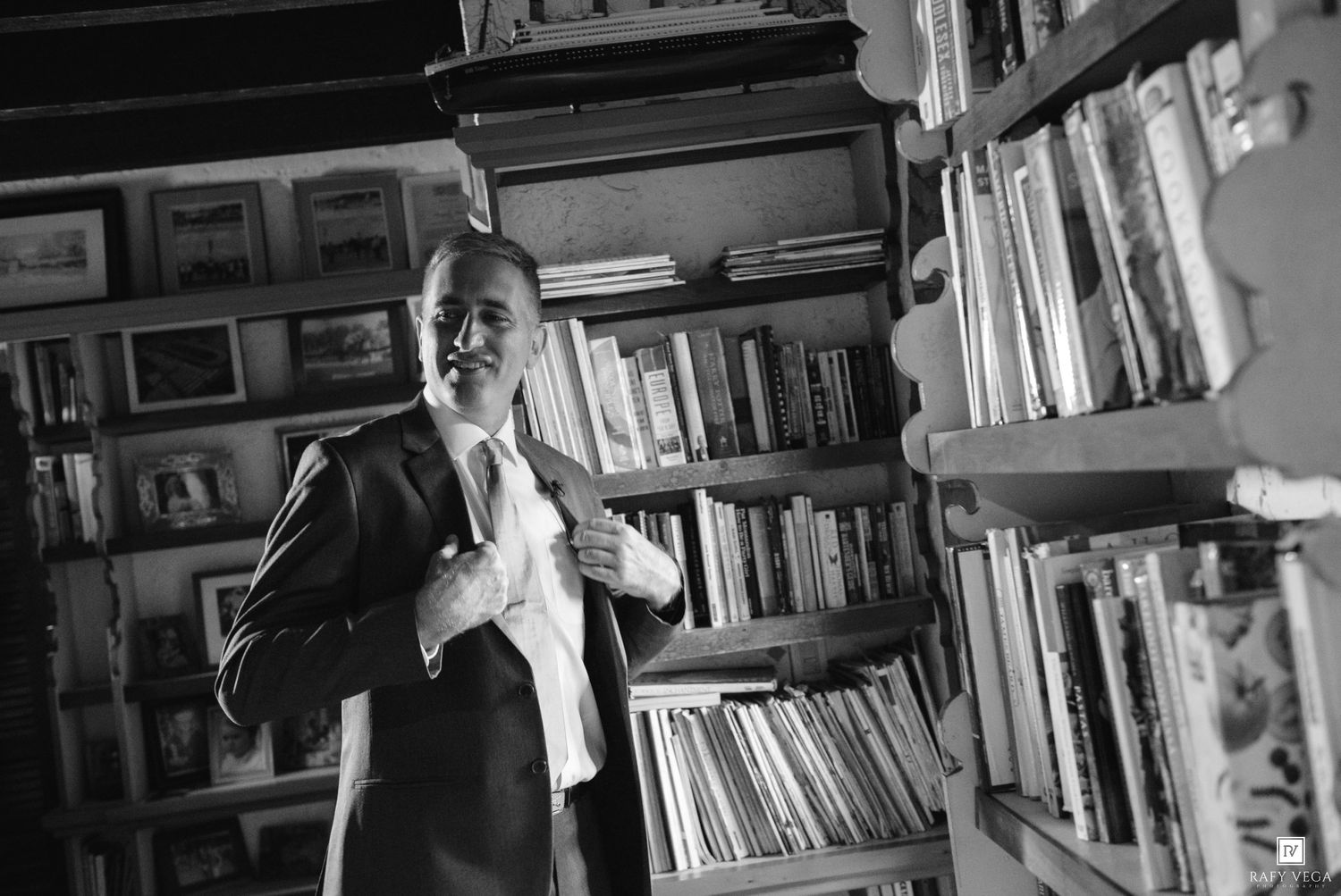 Black and white photo of a smiling man in a suit standing in front of bookshelves filled with books.