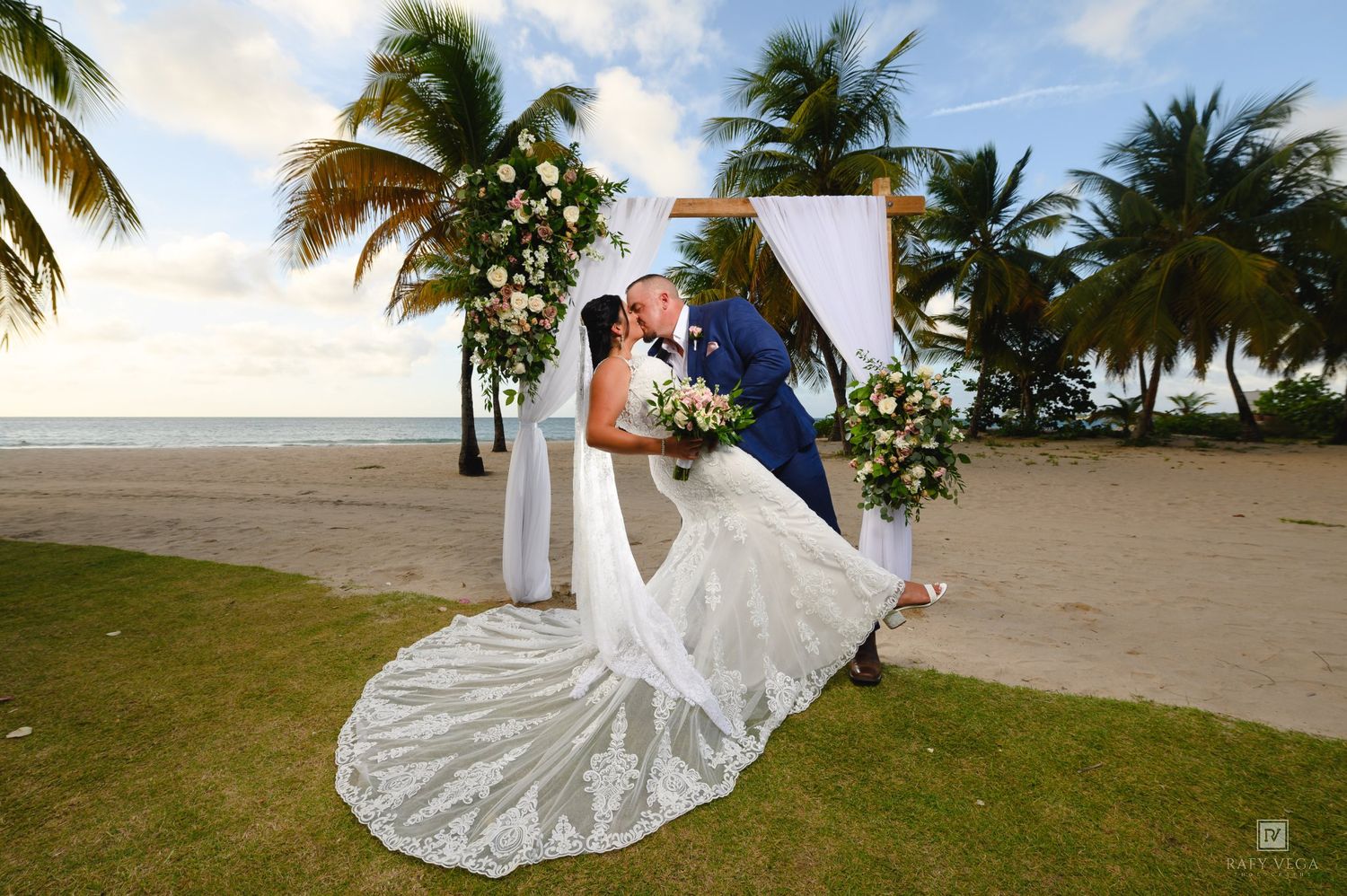 Couple sharing a romantic kiss at a tropical beach wedding ceremony with palm trees and a floral arch backdrop.