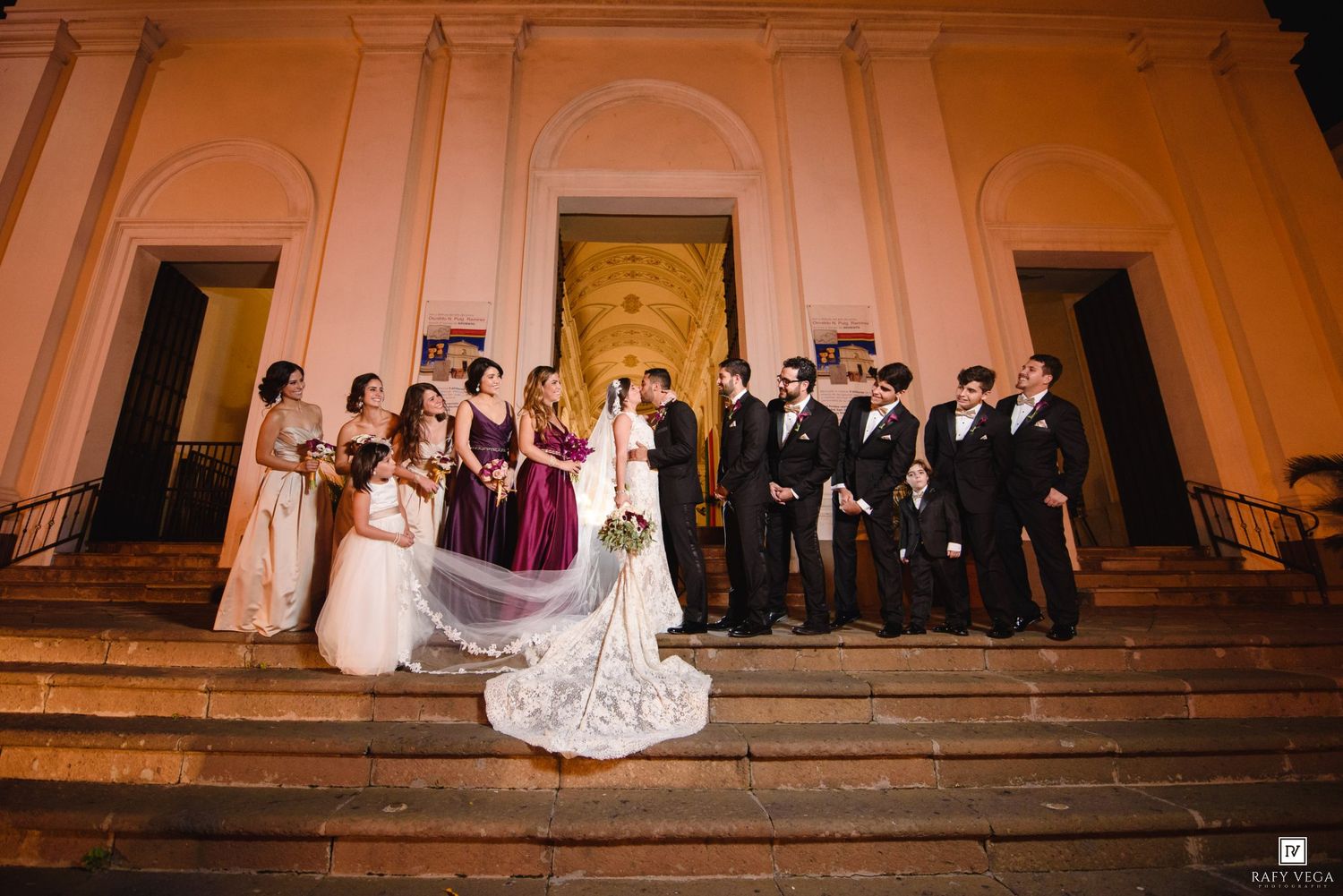 Wedding party poses on grand church steps at night, bride in lace gown with long veil, bridesmaids in burgundy, groomsmen in black.