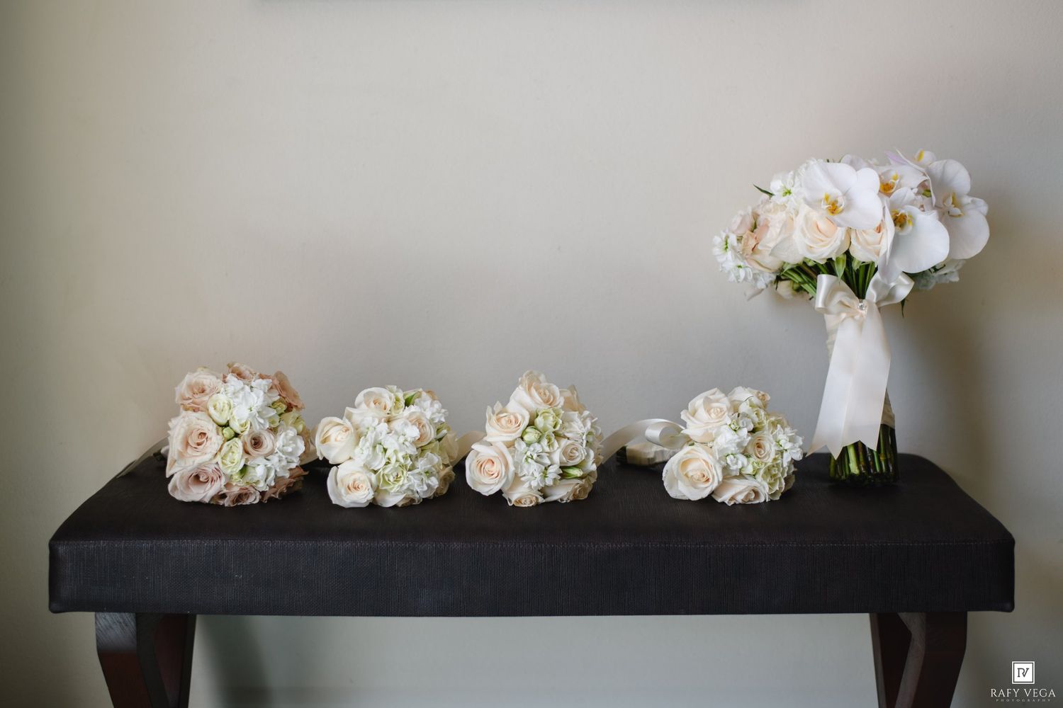 Four white and blush bridal bouquets arranged on a dark bench, with an elegant white orchid bouquet on the right.