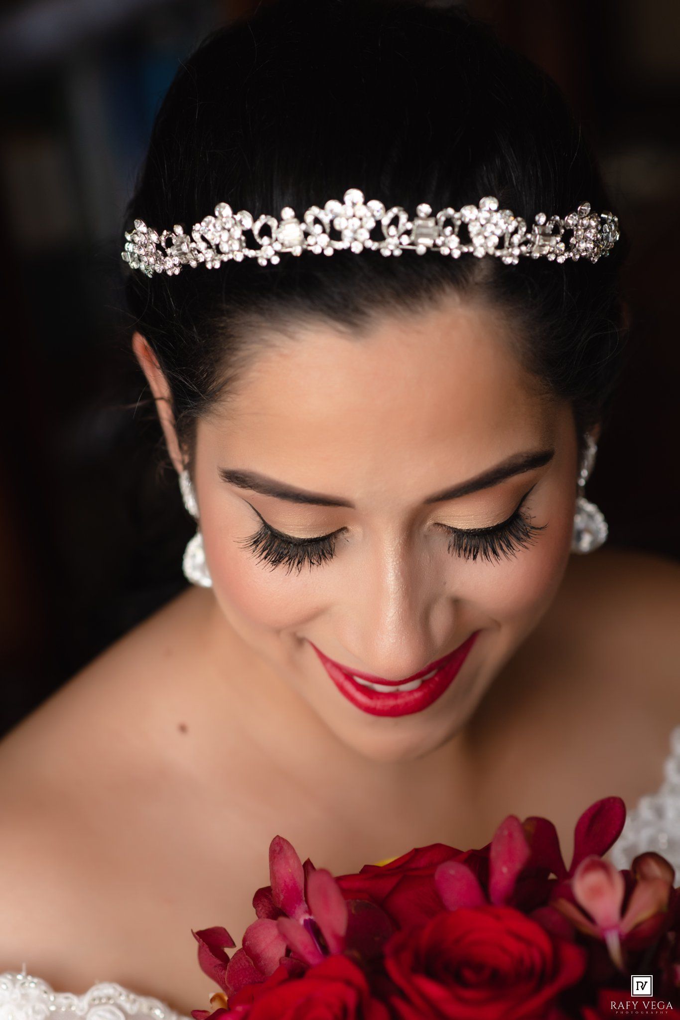 A beautiful bride wearing a pearl tiara and red lipstick holds a bouquet of red flowers.