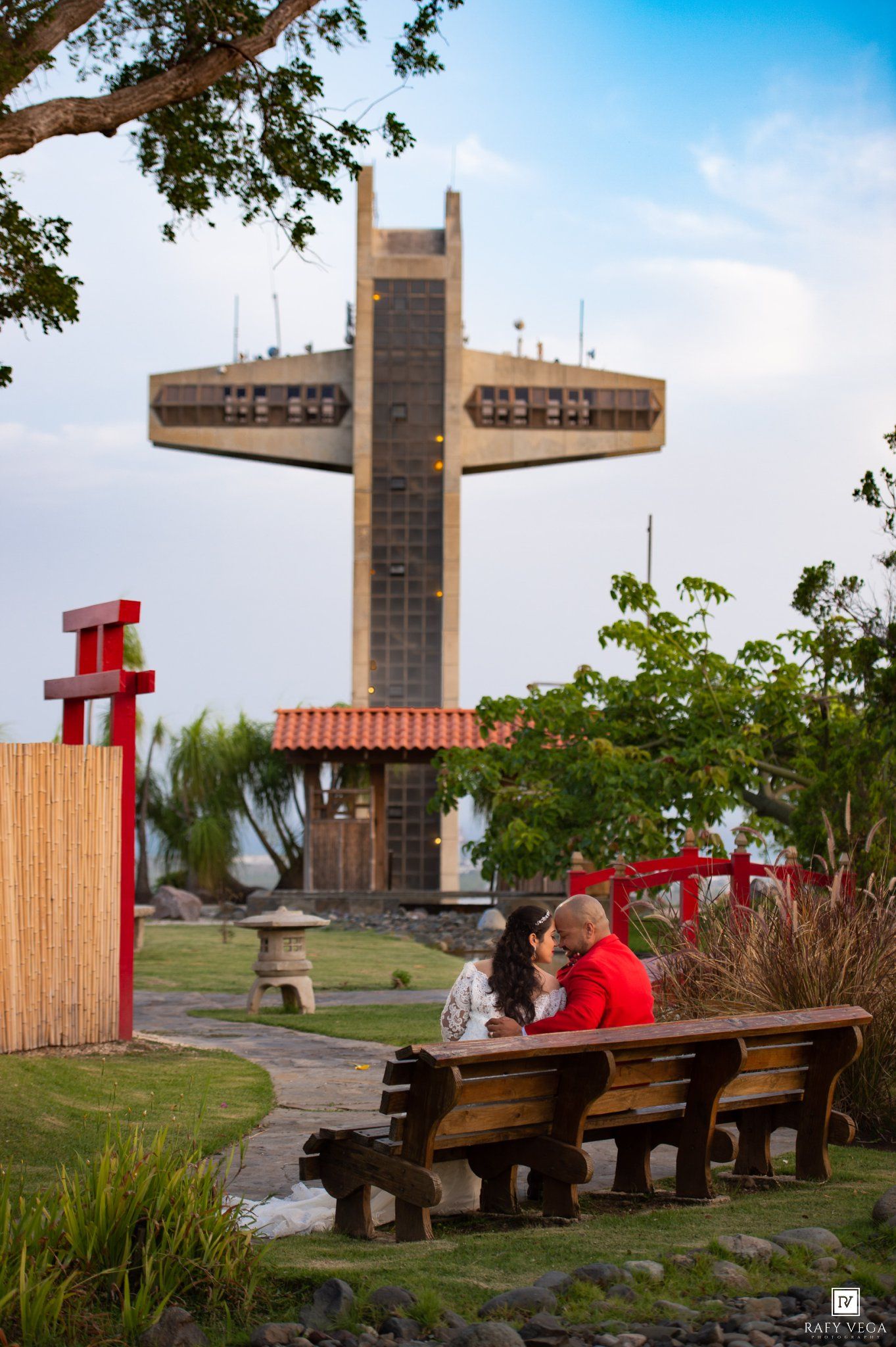 Couple sharing a romantic moment on a wooden bench with the iconic Porta Coeli Cross monument in Ponce, Puerto Rico behind them.