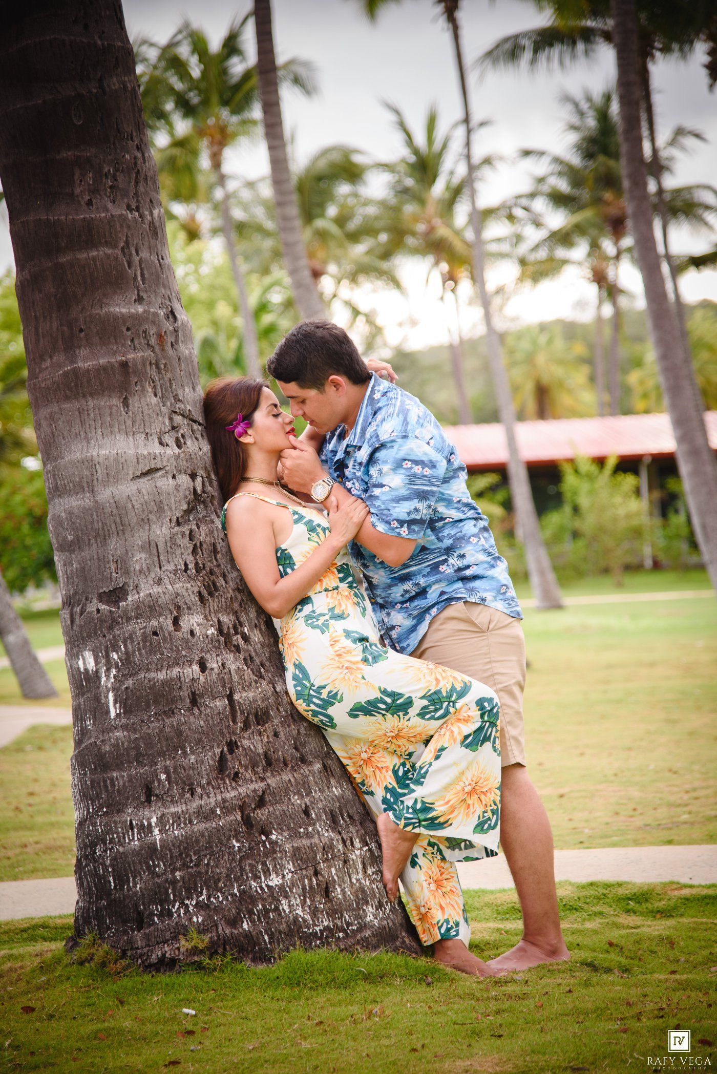 Couple sharing a romantic kiss leaning against a palm tree in a tropical garden, wearing floral outfits.