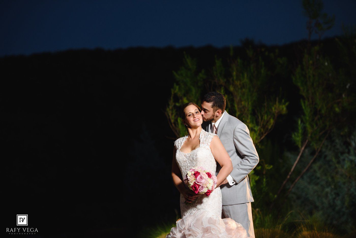 Bride and groom pose intimately at night with bride holding pink bouquet in elegant wedding portrait.