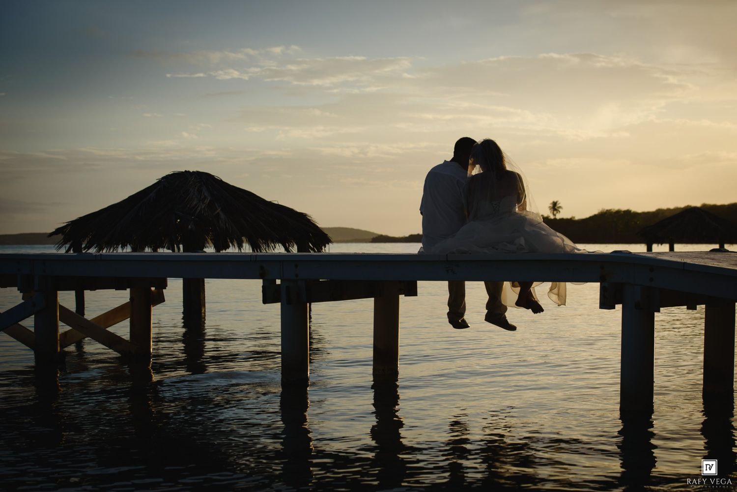 Silhouette of a bride and groom sitting on a dock at sunset over calm tropical waters.