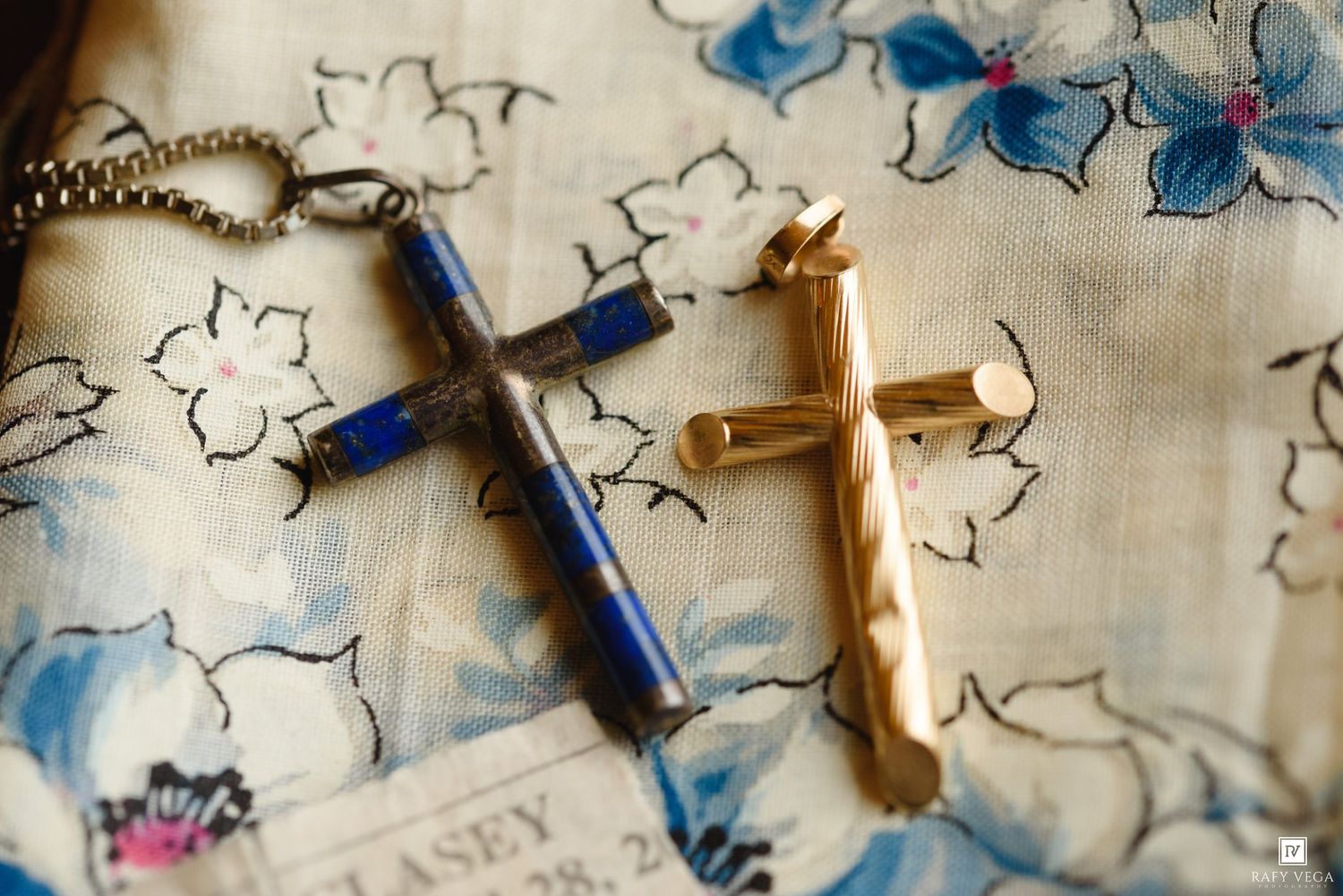Two cross pendants, one blue lapis and silver, one gold, resting on floral fabric.