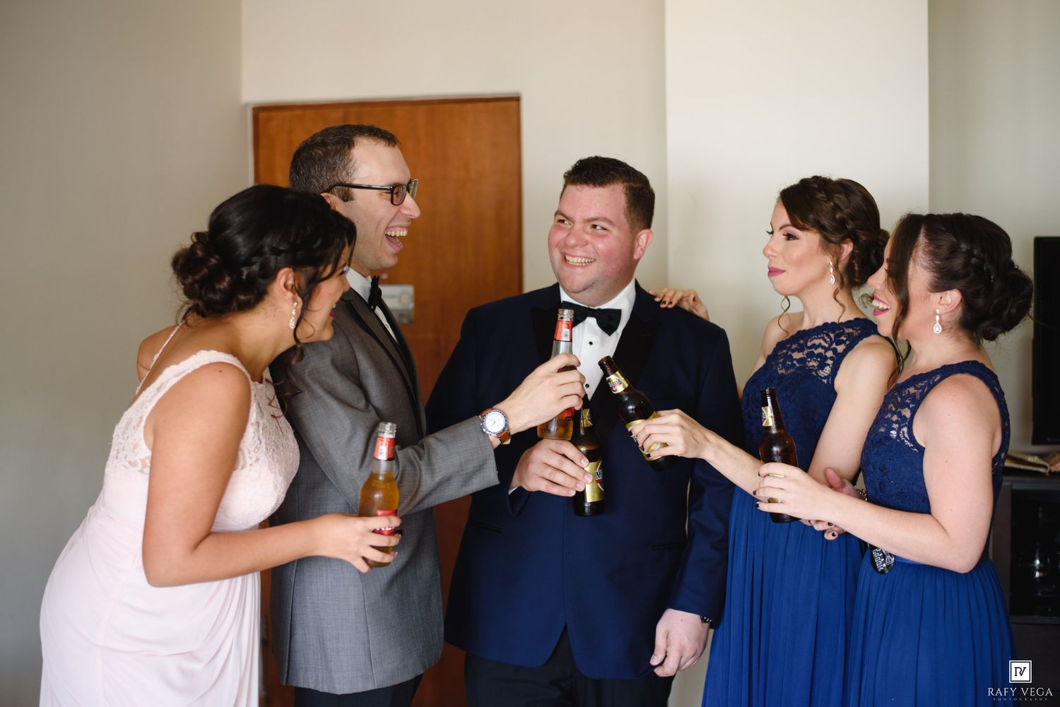 Wedding party in formal attire toasting with drinks, bridesmaids in navy blue dresses celebrating with groom.