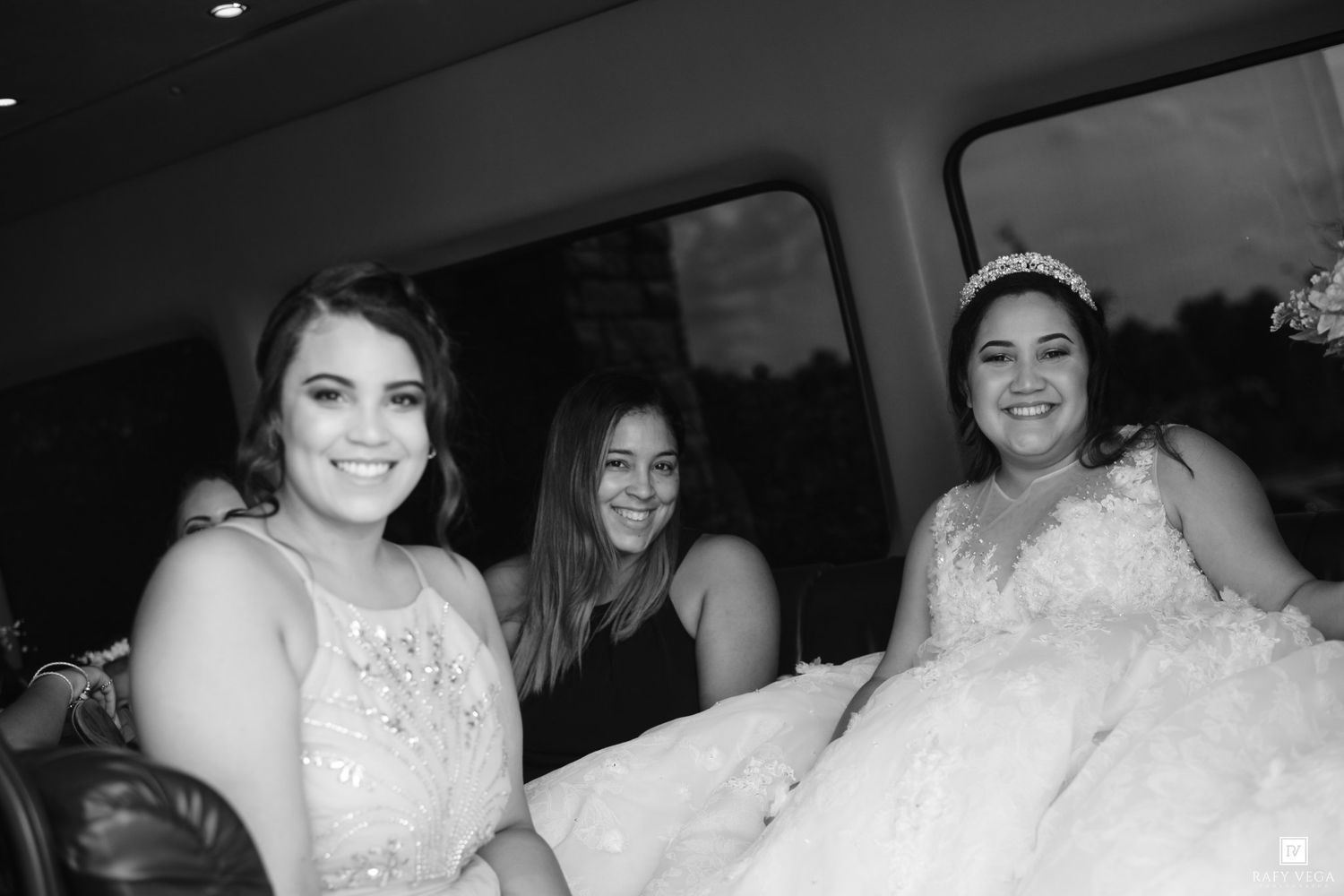 Black and white photo of three smiling women in formal attire, including a bride in a lace gown, seated in a limousine.