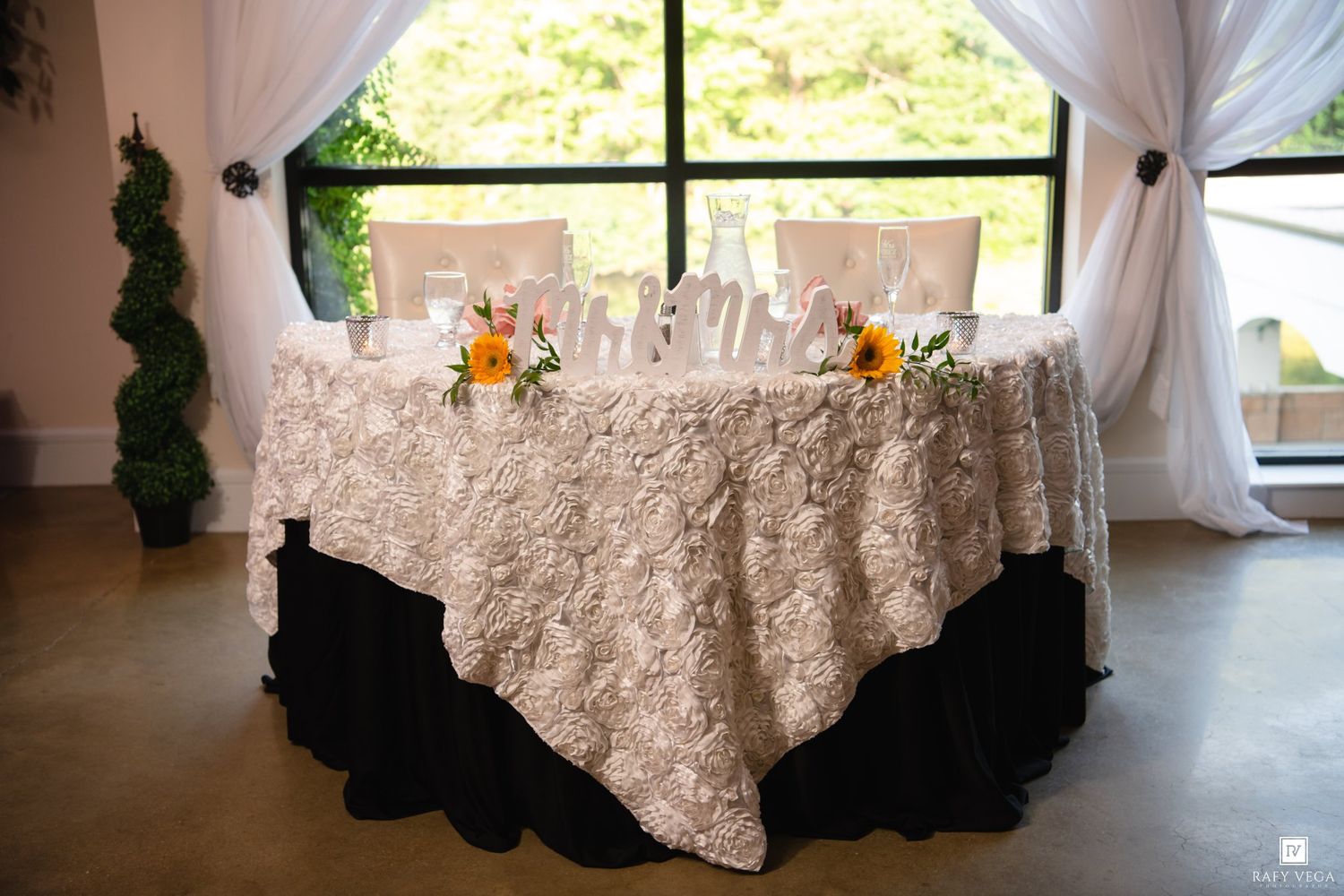 Sweetheart table with ivory rosette lace overlay, Mr. and Mrs. signs, and orange floral centerpiece near draped windows.