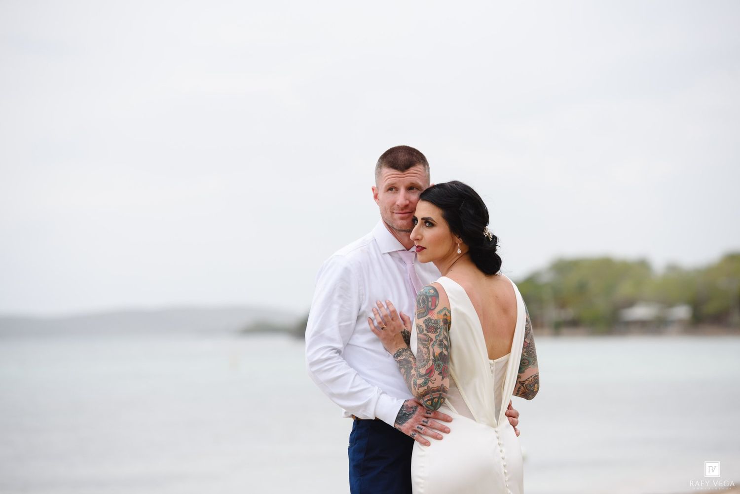 Tattooed groom stands behind bride on beach, bride in sleek white backless gown, both gazing into the distance.