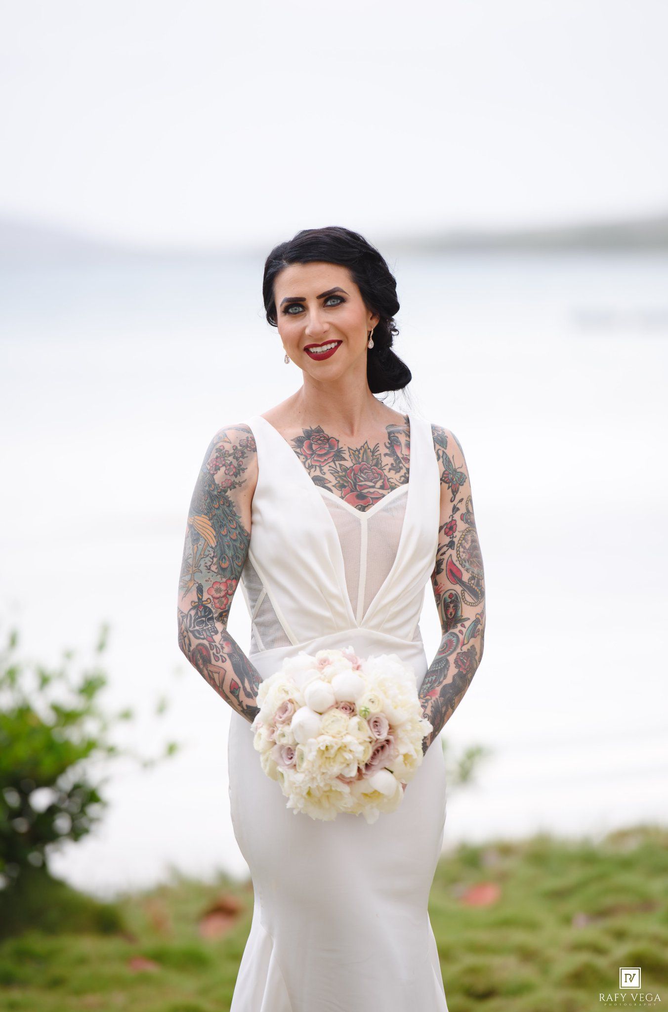 Tattooed bride in white fitted gown holding white floral bouquet, smiling outdoors near waterfront on wedding day.