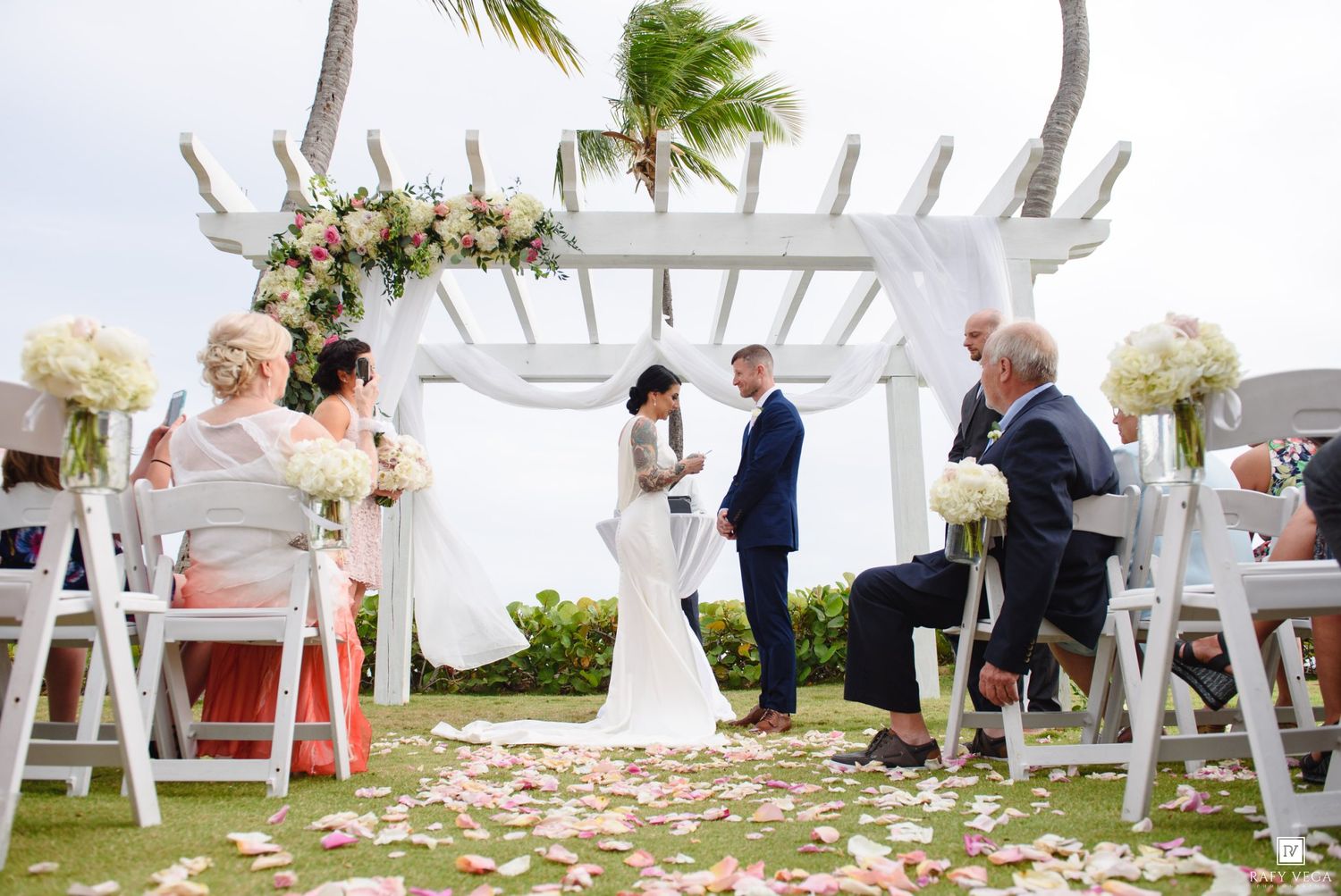 Outdoor beach wedding ceremony under white pergola with floral arch, rose petals on grass, bride and groom exchanging vows.