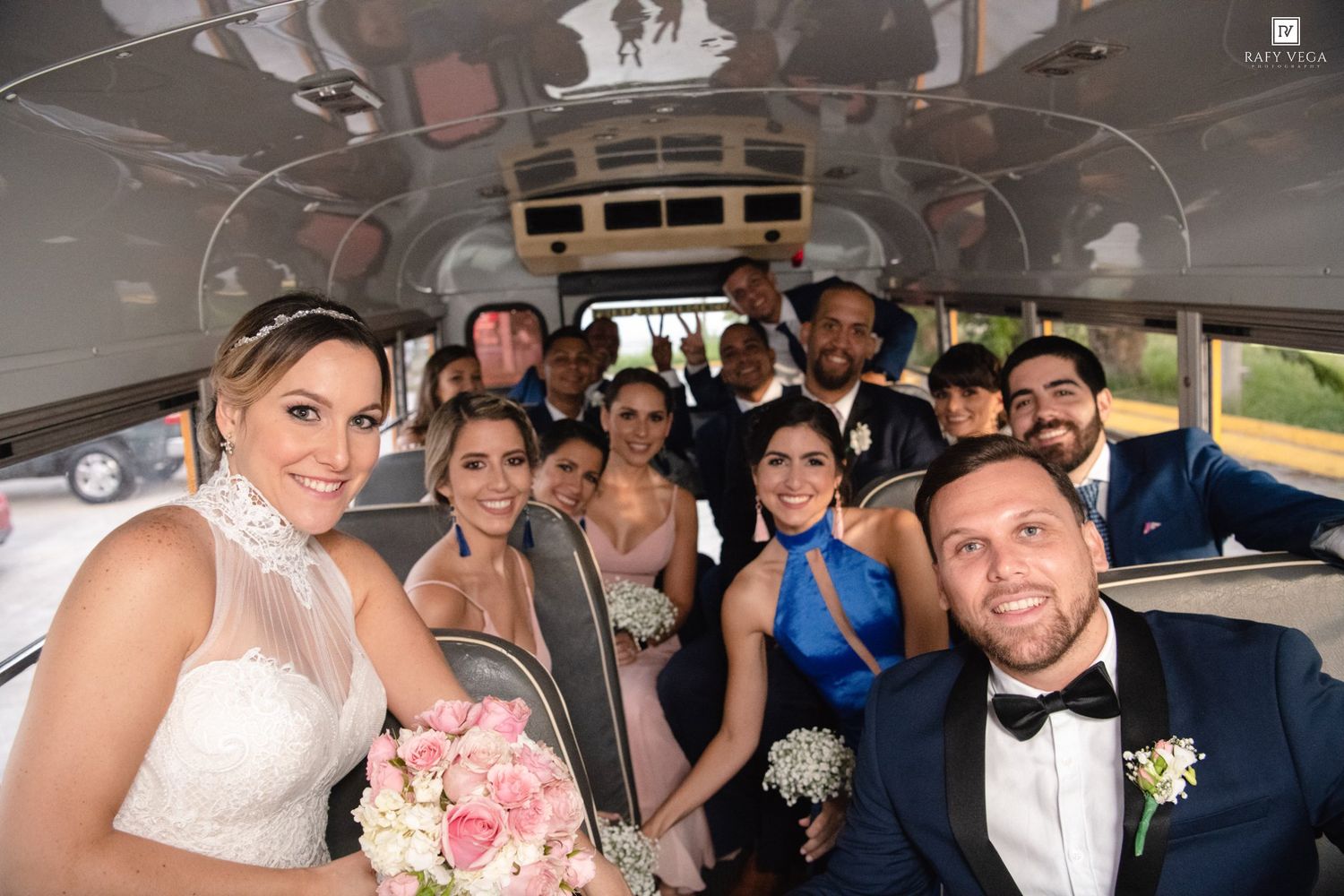 Wedding party smiling inside a decorated party bus, bride holding pink bouquet with groom in tuxedo surrounded by bridesmaids and groomsmen.