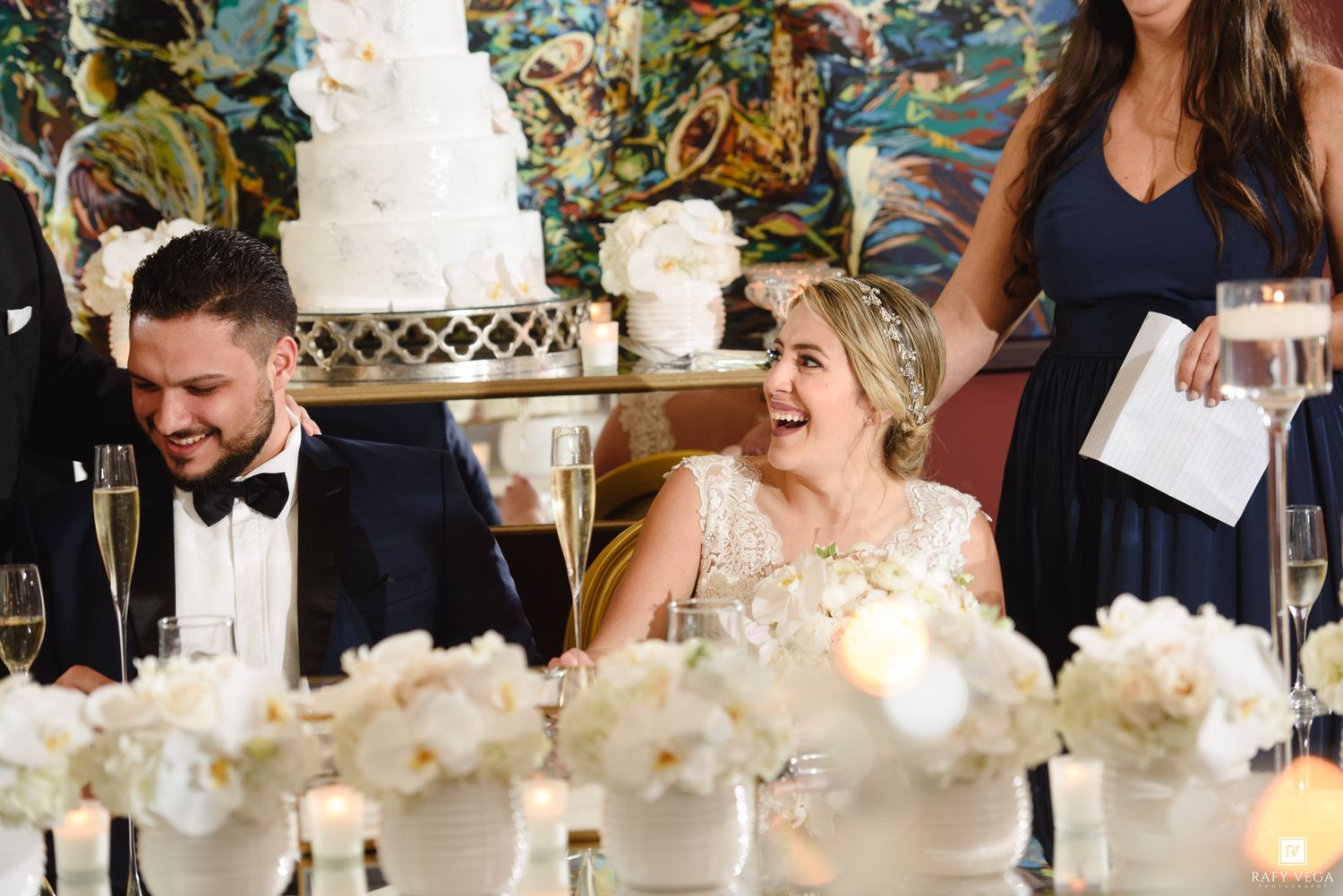 Bride and groom laughing at wedding reception table with white floral centerpieces and tiered cake in background.