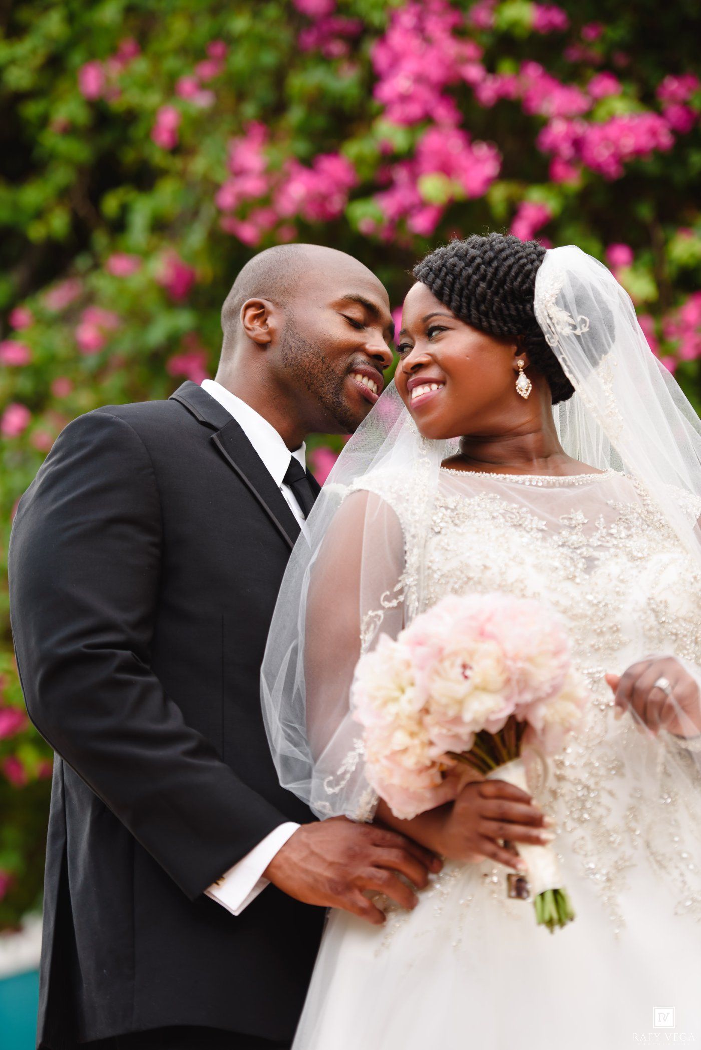 A smiling bride in a lace gown and veil holds a pink bouquet as the groom in a black suit leans in close outdoors.