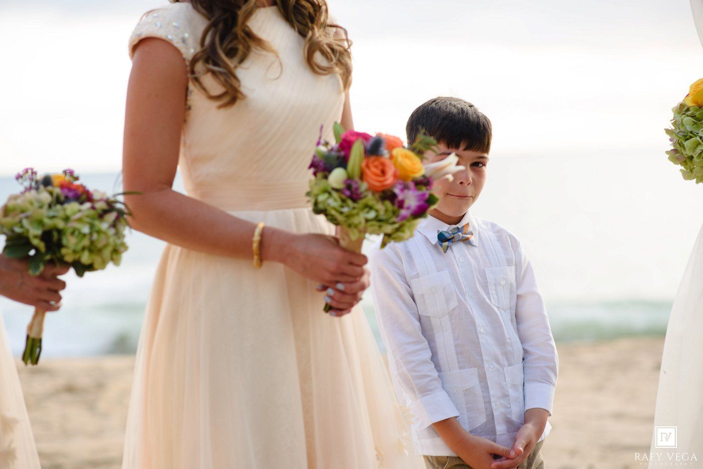 Young ring bearer in bow tie stands beside bridesmaid holding colorful bouquet at beach wedding ceremony.