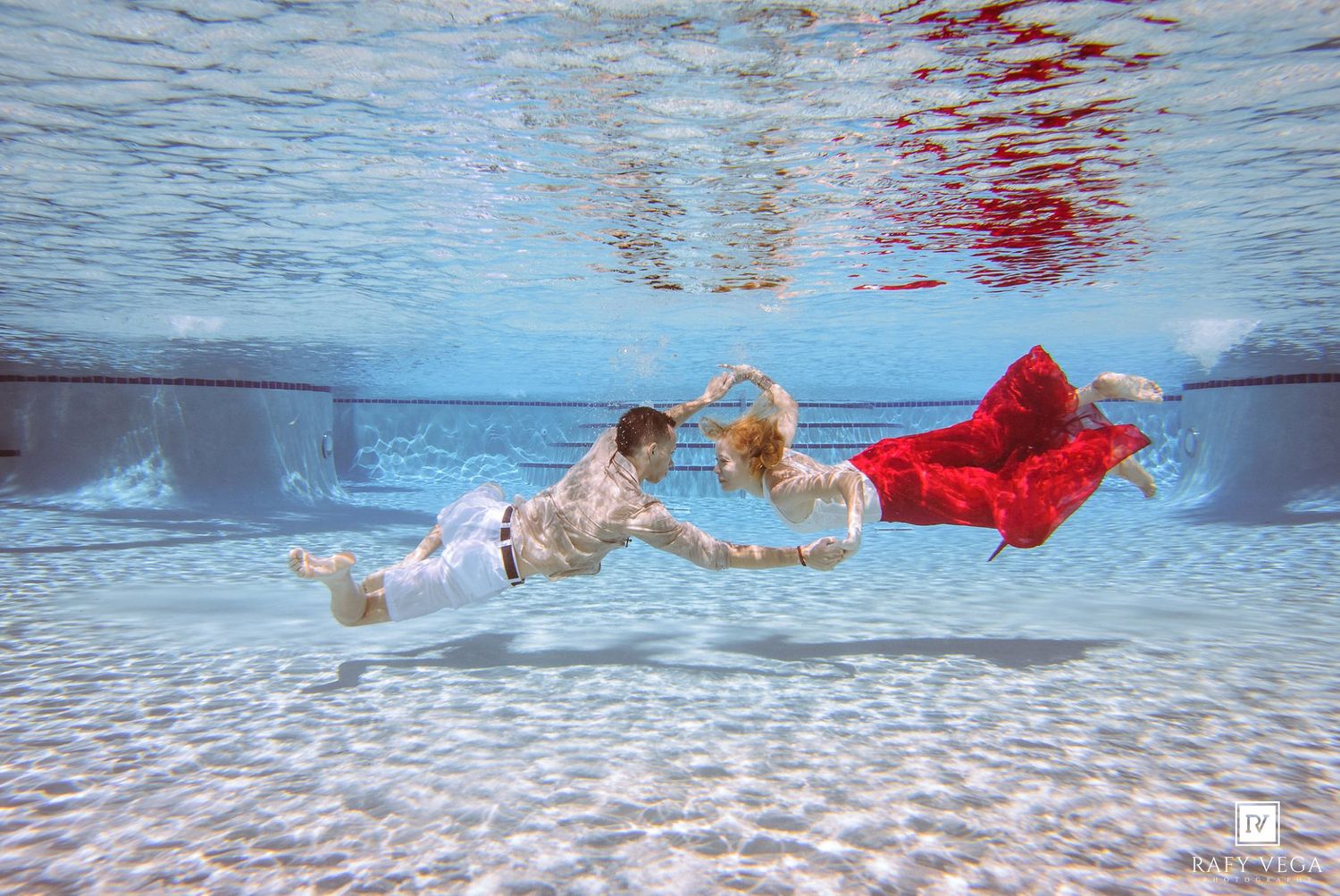 Couple in elegant red and white attire posing romantically underwater in a clear blue swimming pool.