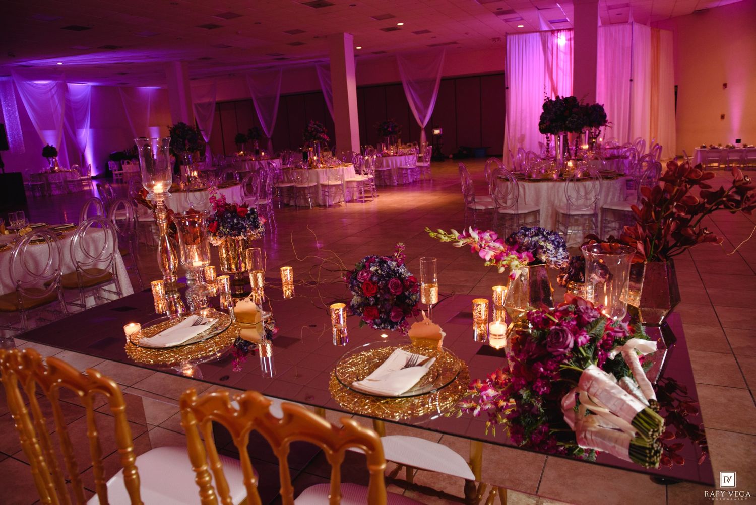 Elegant wedding reception ballroom with purple uplighting, gold chairs, floral centerpieces, and candlelit mirror tables.