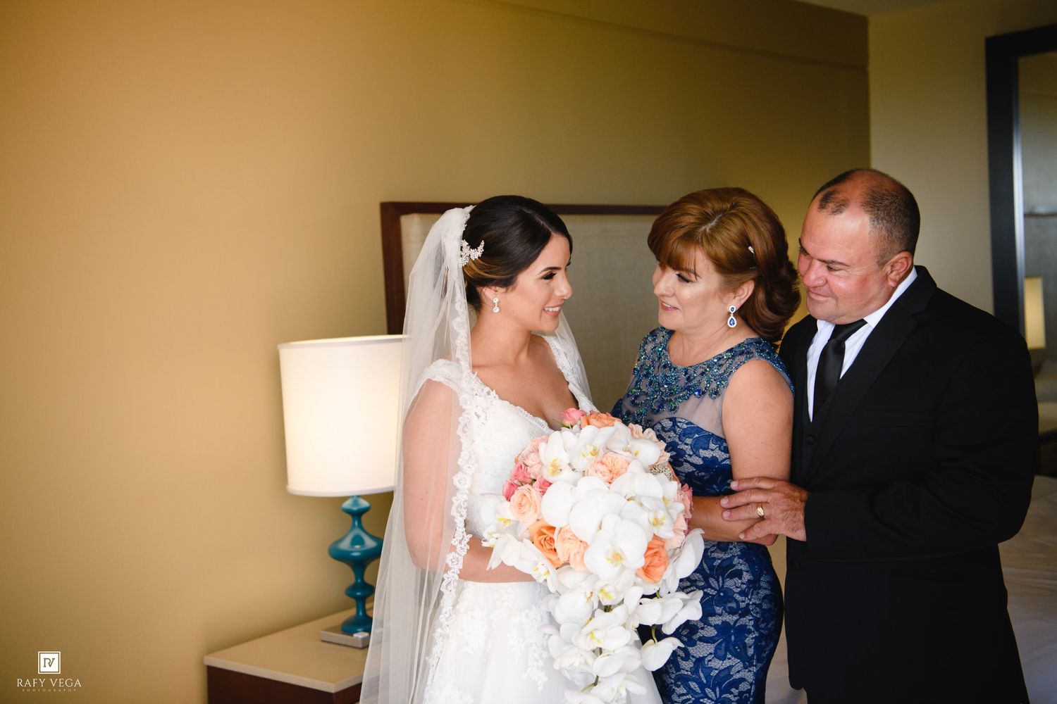 Bride in white lace gown shares emotional moment with parents before wedding ceremony indoors.