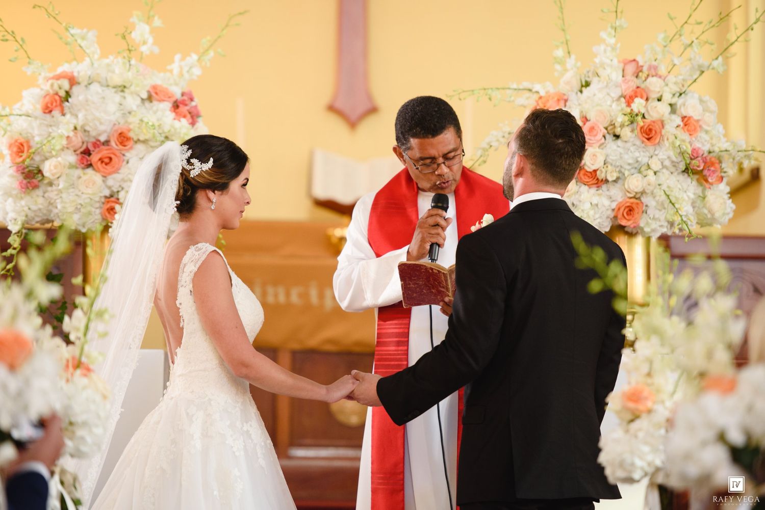 Bride and groom holding hands during church wedding ceremony while priest officiates at flower-adorned altar.