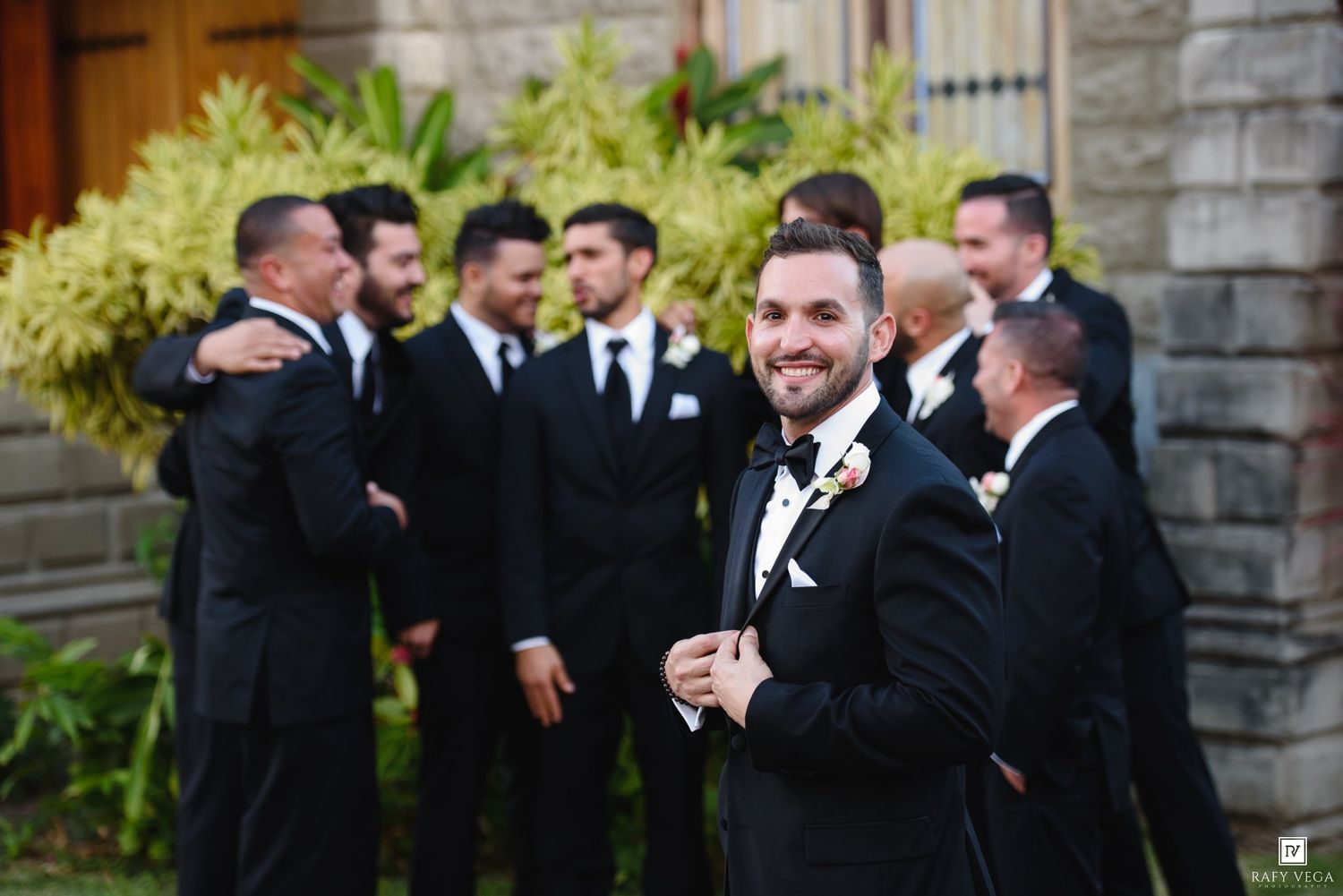 Groom smiling in black tuxedo with groomsmen socializing outside stone building at wedding.