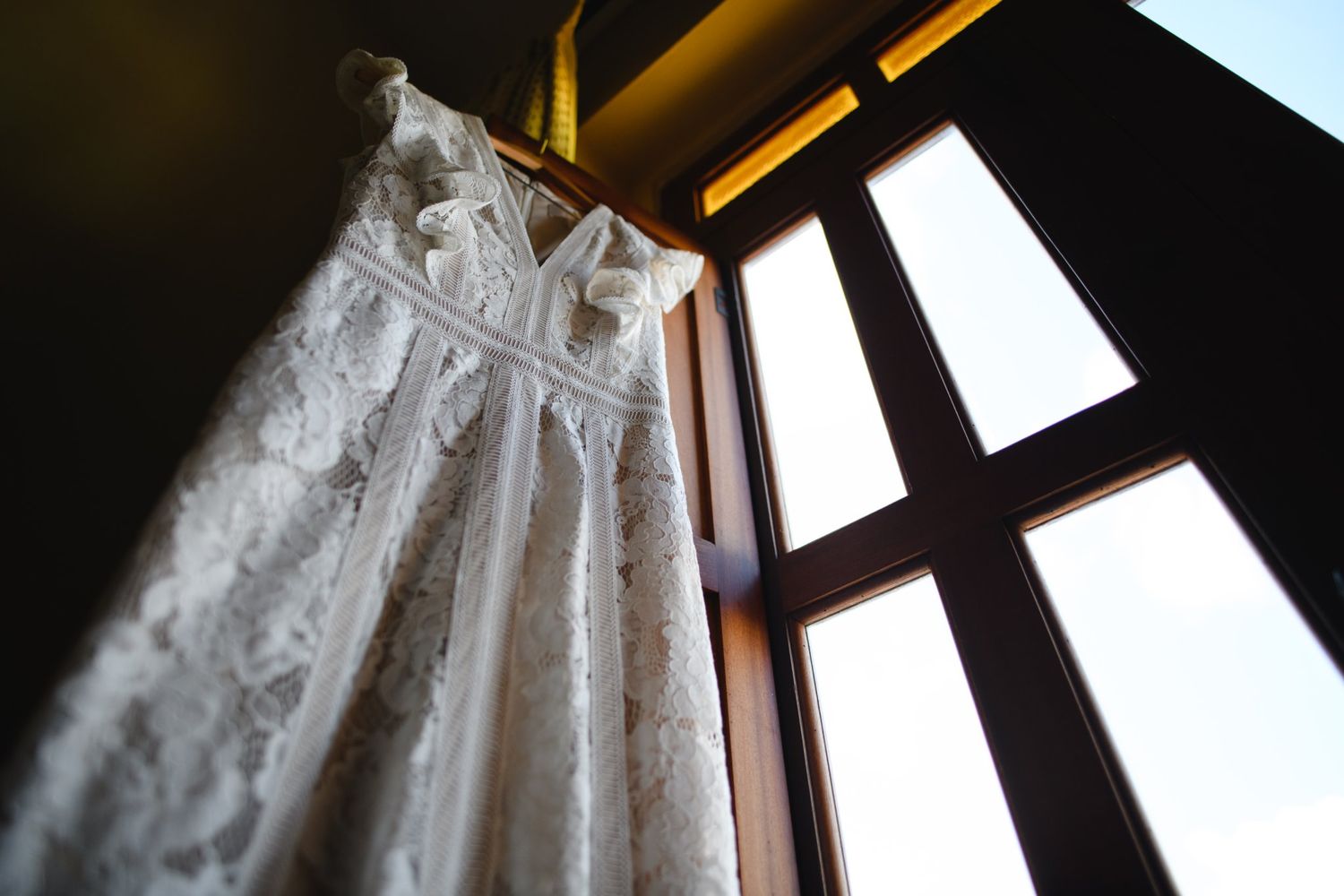 Elegant white lace wedding dress hanging on a hanger near a sunlit window with wooden frames.