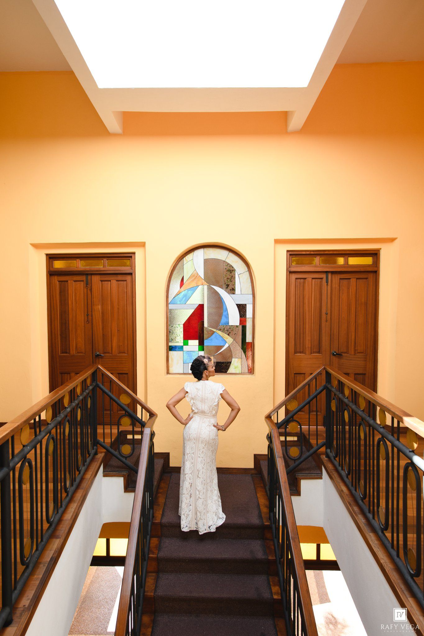 Bride in white lace gown stands on staircase landing before colorful stained glass artwork in elegant venue.