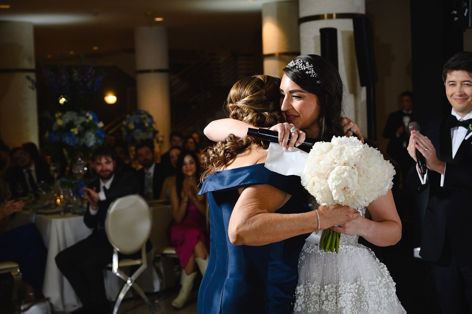 Bride in white gown hugging a guest in navy dress while holding white bouquet at elegant wedding reception.
