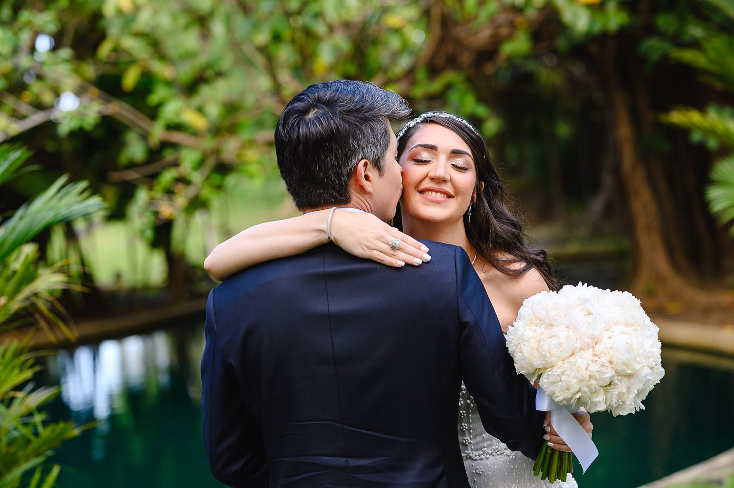 Smiling bride holds white hydrangea bouquet as groom kisses her cheek in a tropical garden wedding setting.