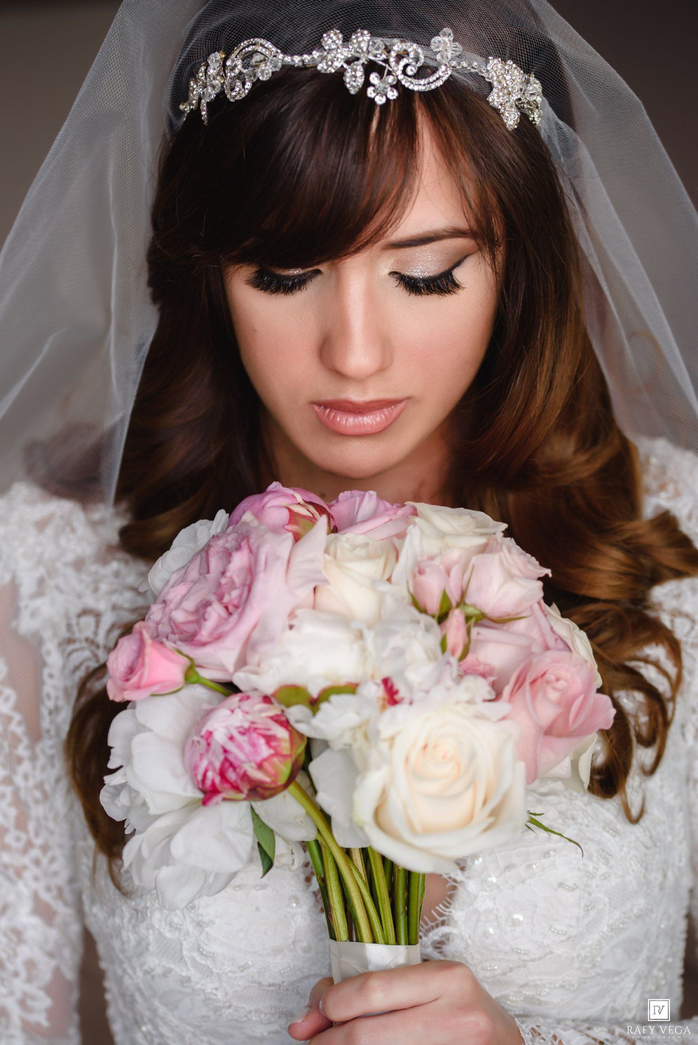 Bride in lace gown wearing crystal tiara and veil holds pink and white rose bouquet, looking down elegantly.