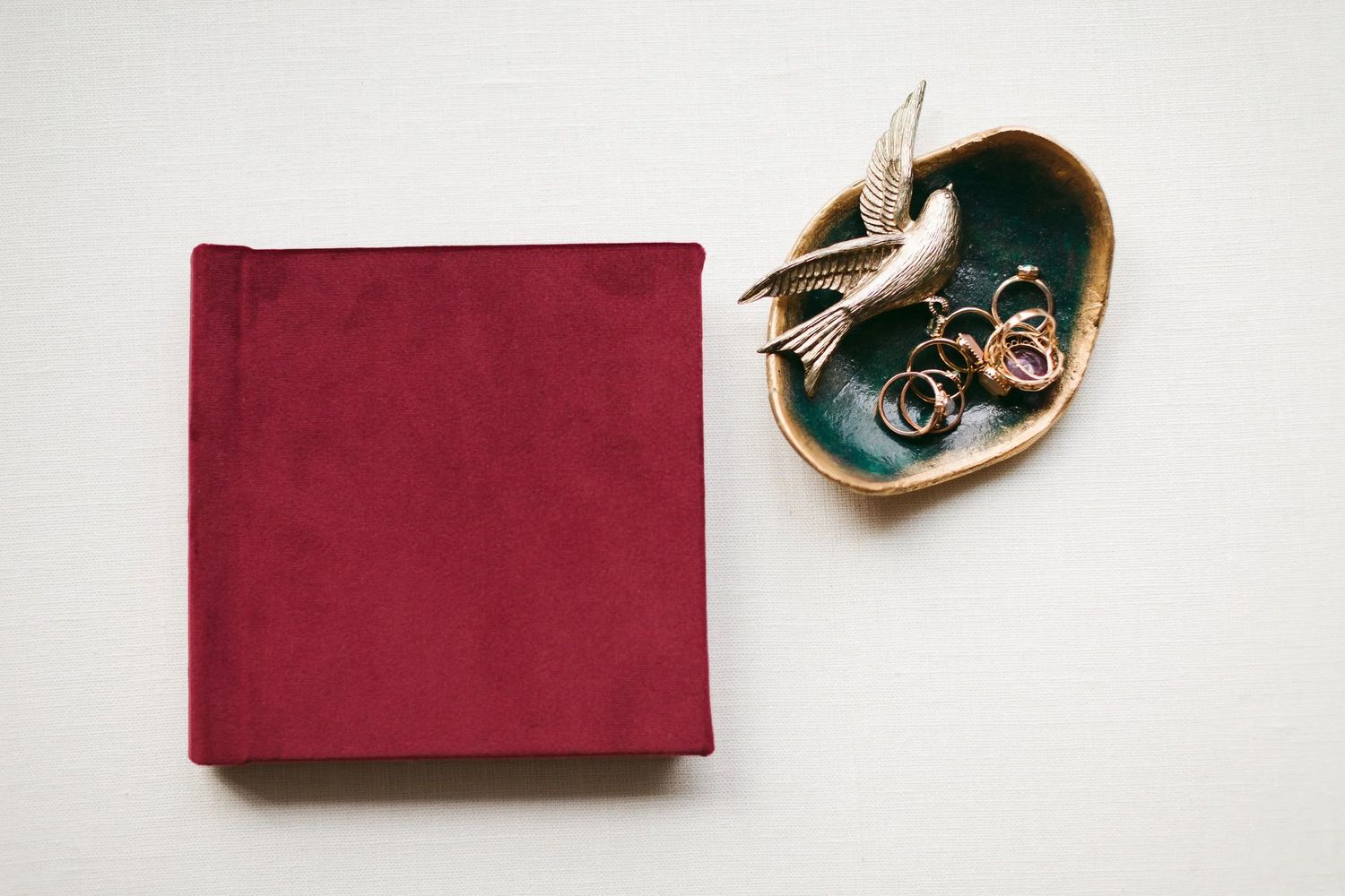 Burgundy velvet hardcover photo album beside a teal ceramic bird dish holding gold jewelry and a brooch.