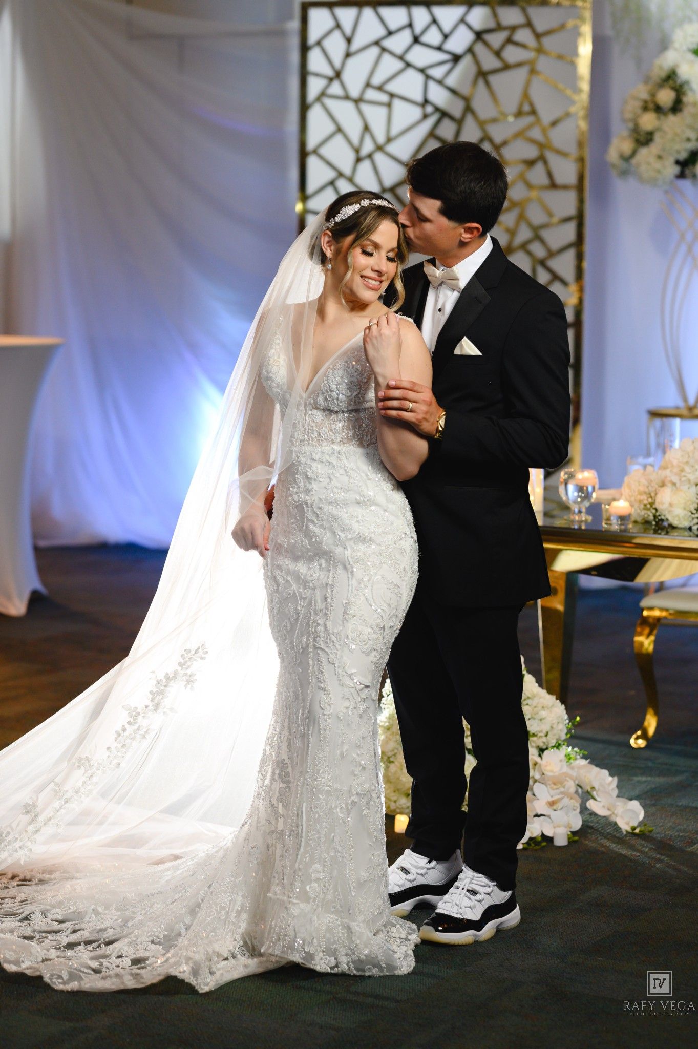Groom in black tuxedo kisses bride in lace mermaid dress and veil during romantic wedding reception moment.
