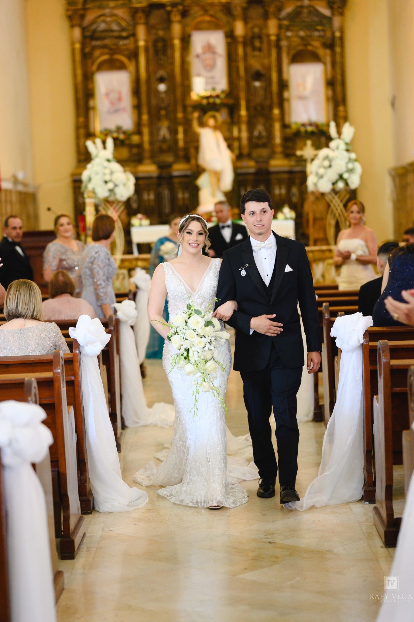 Newlyweds walk down the church aisle smiling, bride in beaded gown holding white bouquet, groom in dark tuxedo.