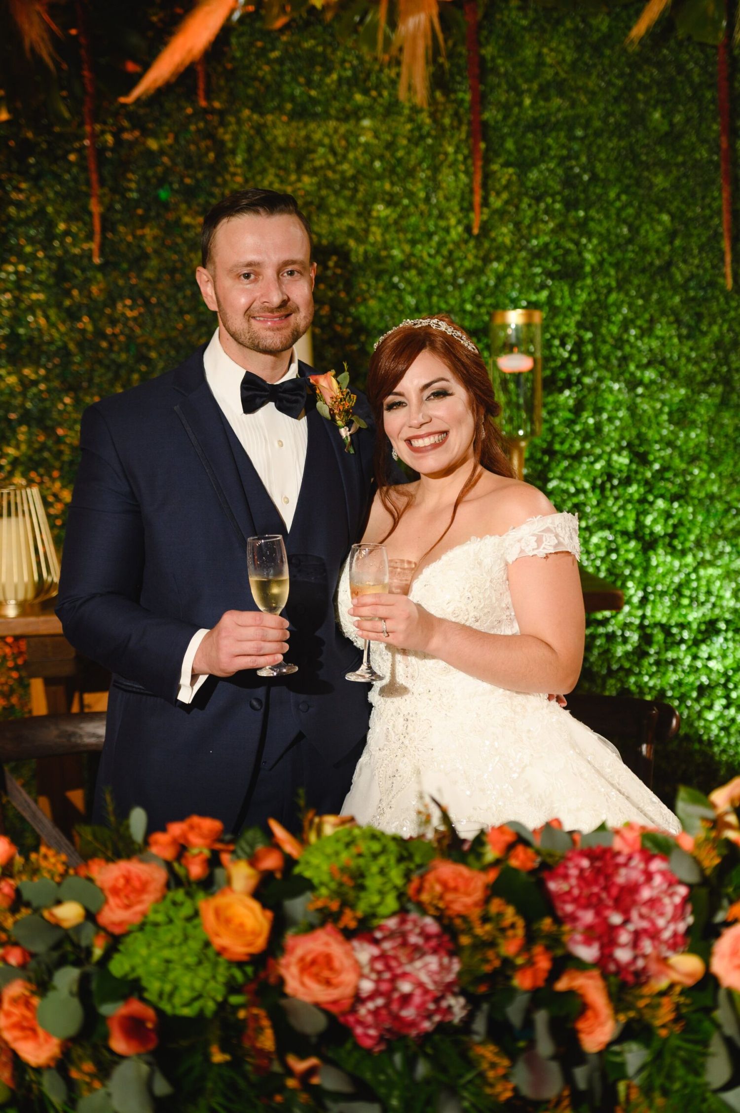 Bride and groom toasting with champagne glasses at wedding reception with floral arrangements and green wall backdrop.