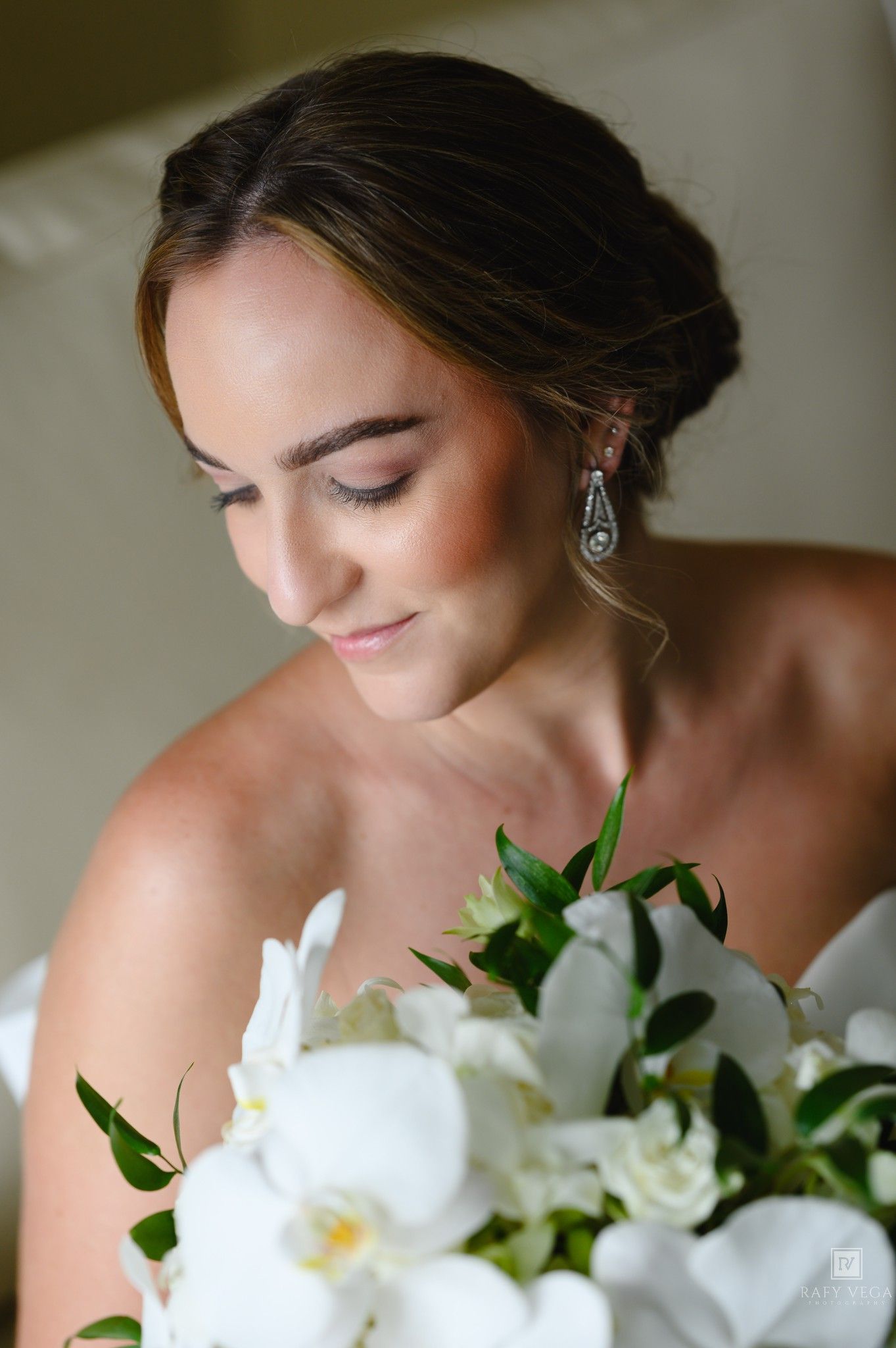 Bride with updo hairstyle and elegant drop earrings gazes down at white orchid bouquet on her wedding day.