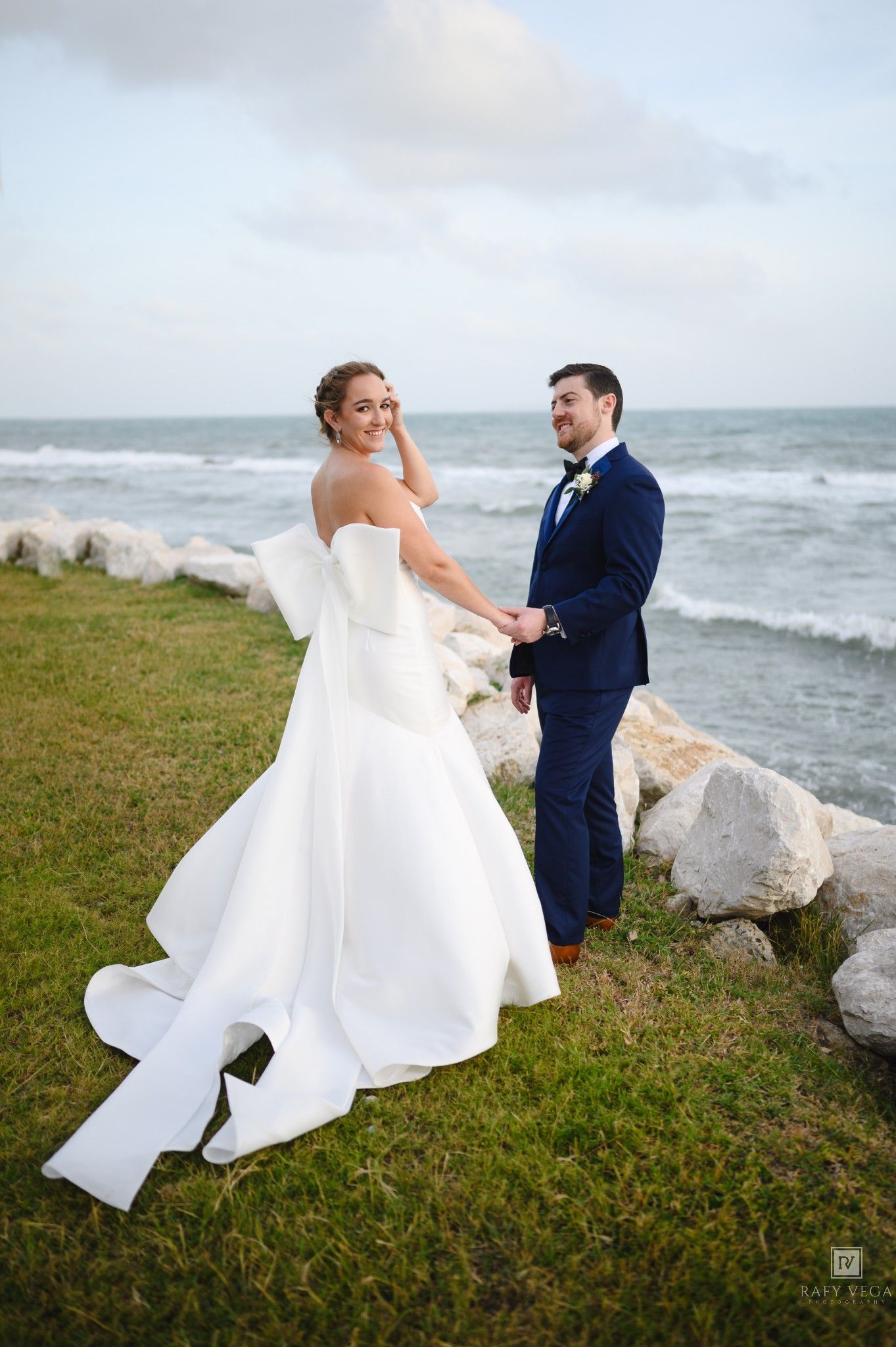 Bride in white strapless gown and groom in navy suit hold hands by rocky oceanside on their wedding day.